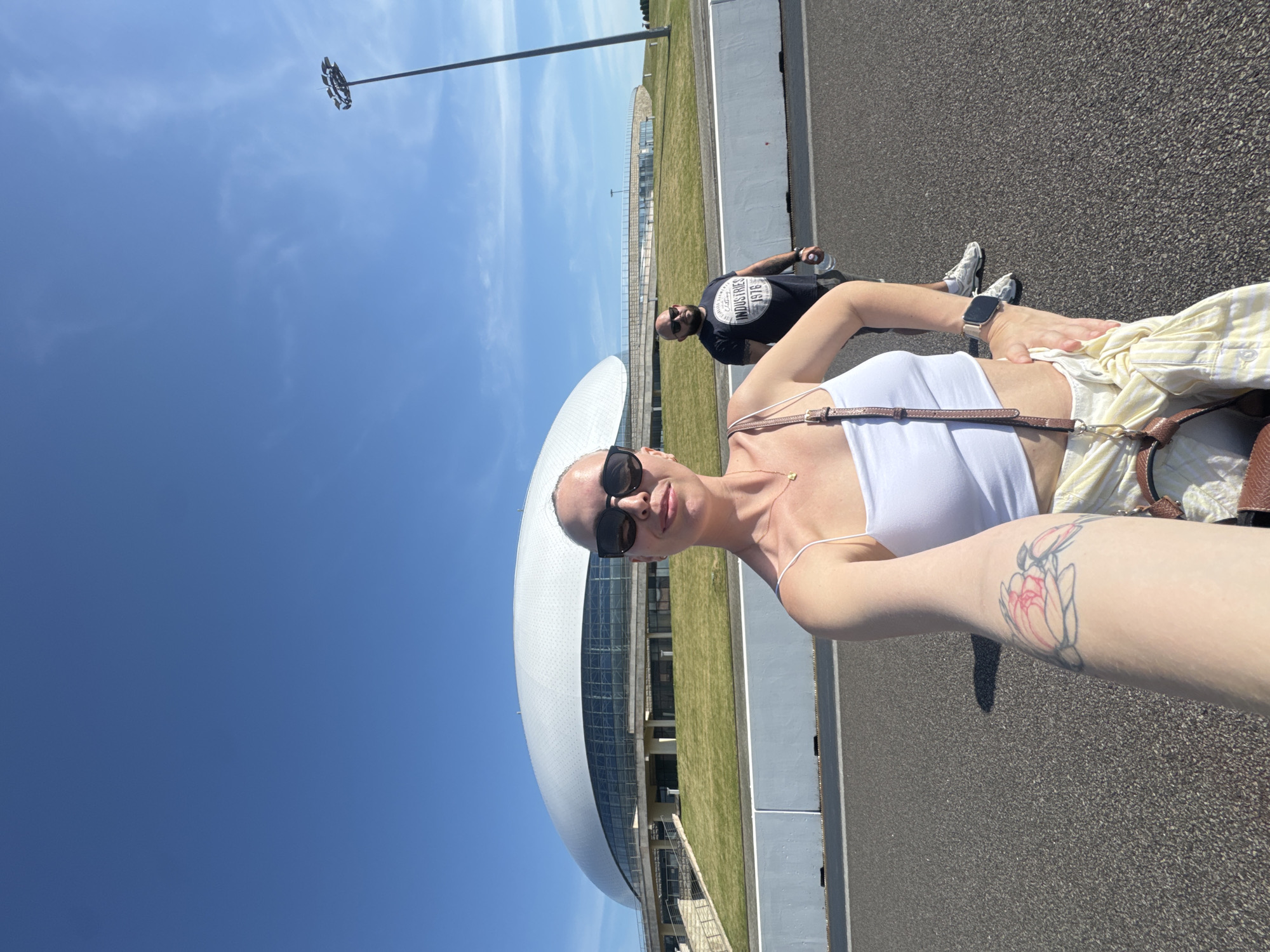 A selfie-style photograph depicts two individuals standing outdoors on a paved surface on a clear, sunny day. In the foreground, a woman with dark sunglasses and a light-colored crop top is visible from the waist up, holding the camera in her extended right arm, which features a floral tattoo. She wears a smartwatch on her left wrist and has a brown shoulder bag strap crossing her chest. Her left hand rests on her hip. Behind her and to the right, a bald man with a beard and dark sunglasses stands, holding a clear plastic water bottle in his right hand. He wears a dark t-shirt with the text "INDUSTRIES 1976" visible and light-colored sneakers. In the background, prominently featured, is the Bolshoy Ice Dome in Sirius, Russia, a large, white, domed building with extensive glass facades on its lower section, situated atop a landscaped green mound. The paved foreground features a white line and low, light-colored concrete barriers. A tall floodlight pole extends into the clear blue sky on the far right.
