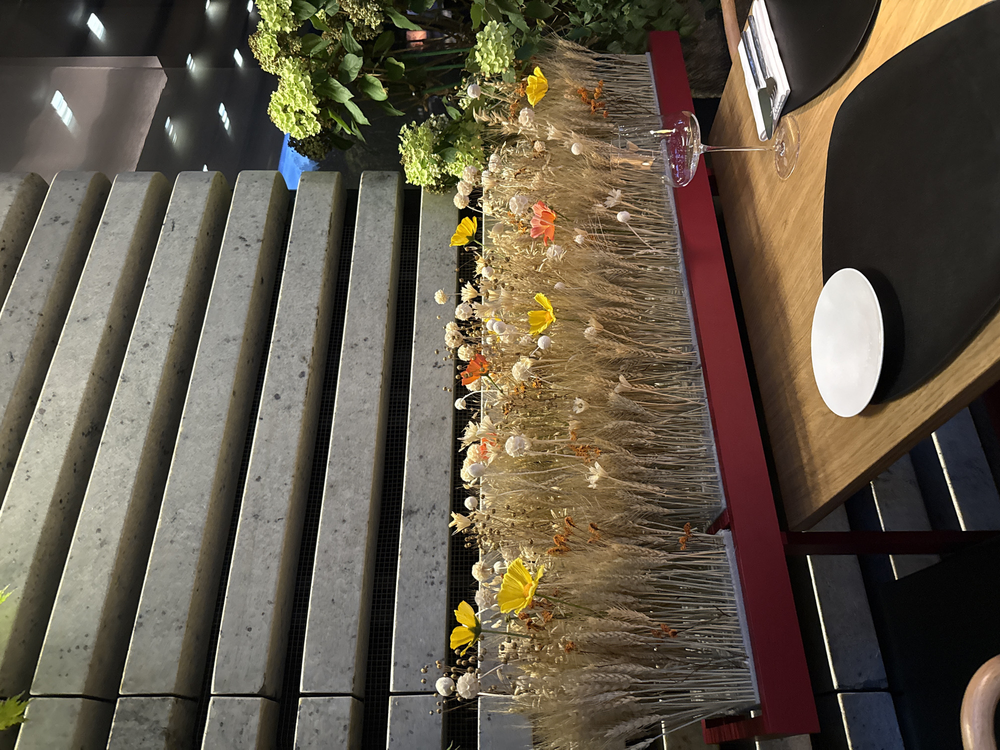 The image depicts a table setting at a restaurant in Moscow, Russia. The table is made of wood and is set with a white plate, a black placemat, and cutlery on a white napkin. A wine glass is placed to the right of the plate, partially filled with red liquid and reflecting the overhead lights. Behind the table, there is a floral arrangement in a red planter, featuring dried wheat stalks, small white and beige dried flowers, and a few colorful faux flowers in yellow and orange. A textured concrete wall with horizontal lines forms the background, with some greenery and artificial lighting visible above and to the right of the floral arrangement. The overall ambiance suggests an indoor dining setting, possibly in the evening.