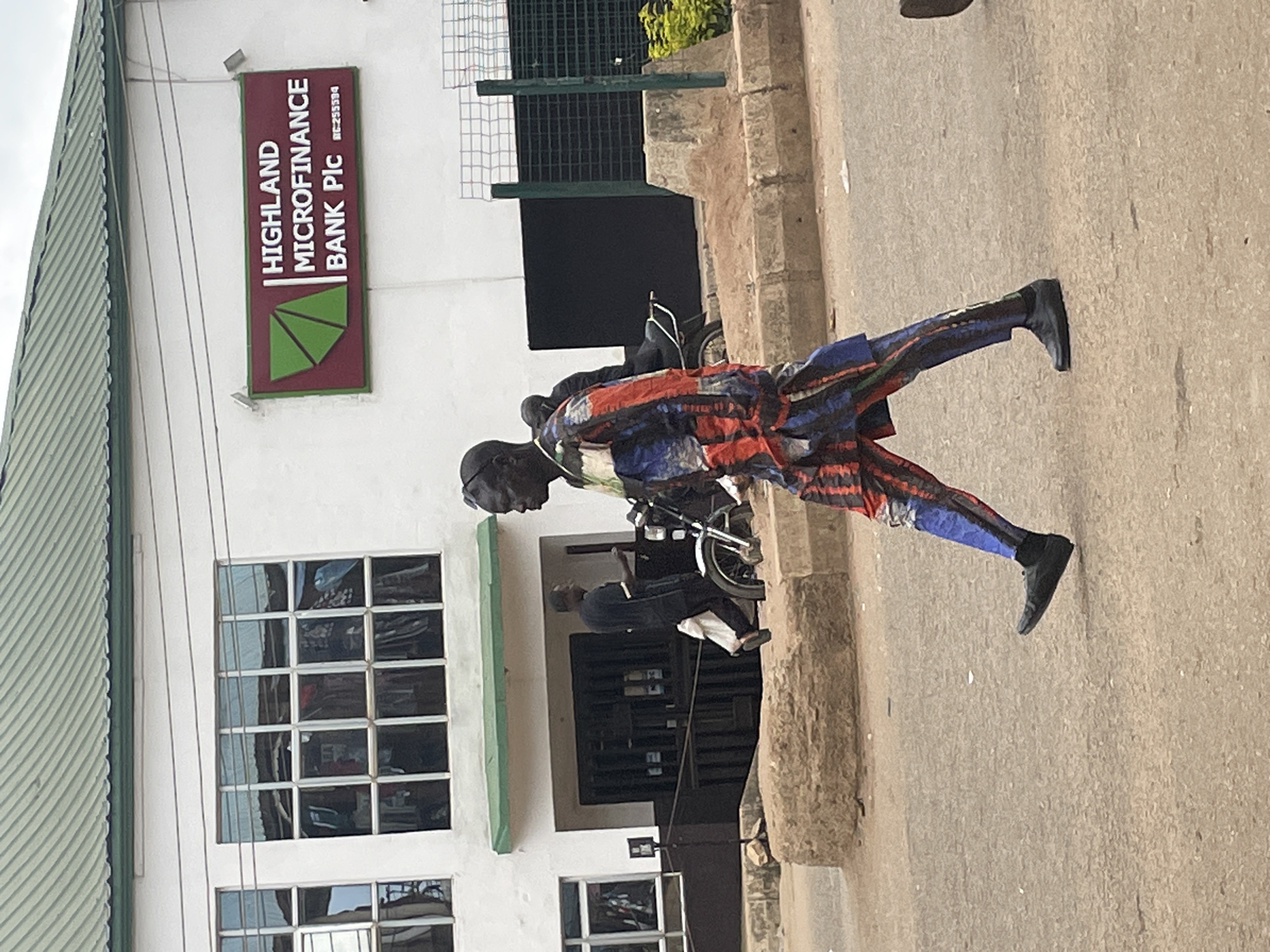 A man in a colorful, patterned outfit walks across a paved street in front of a building with a green roof and white walls. The building has multiple windows and a sign that reads "HIGHLAND MICROFINANCE BANK Plc." Another man, wearing a dark shirt and pants, walks in the background near the entrance of the building, carrying a white bag. A motorcycle is parked behind him. The street is lined with concrete barriers and some vegetation. The scene appears to be in an urban setting in Bukuru, Nigeria.