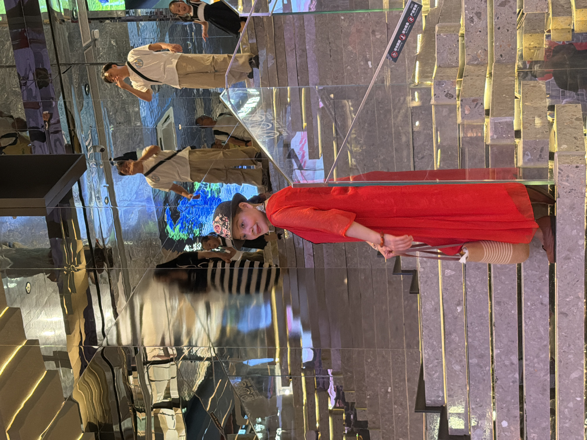 In Yuzhong District, China, a woman stands on a multi-tiered staircase, her red dress flowing down to her feet. She wears a wide-brimmed hat with a floral pattern and large hoop earrings. A straw bag with brown straps hangs from her left arm. The staircase is made of stone with illuminated edges and is surrounded by mirrored walls that reflect the surroundings, including other people and a colorful digital display. To the right of the woman, a young girl in a black and white dress is visible on a higher level of the stairs. Several other individuals, dressed in casual attire, are also present on the staircase and in the mirrored reflections. The overall environment appears to be an indoor public space, possibly a mall or entertainment venue, with modern architecture and lighting.