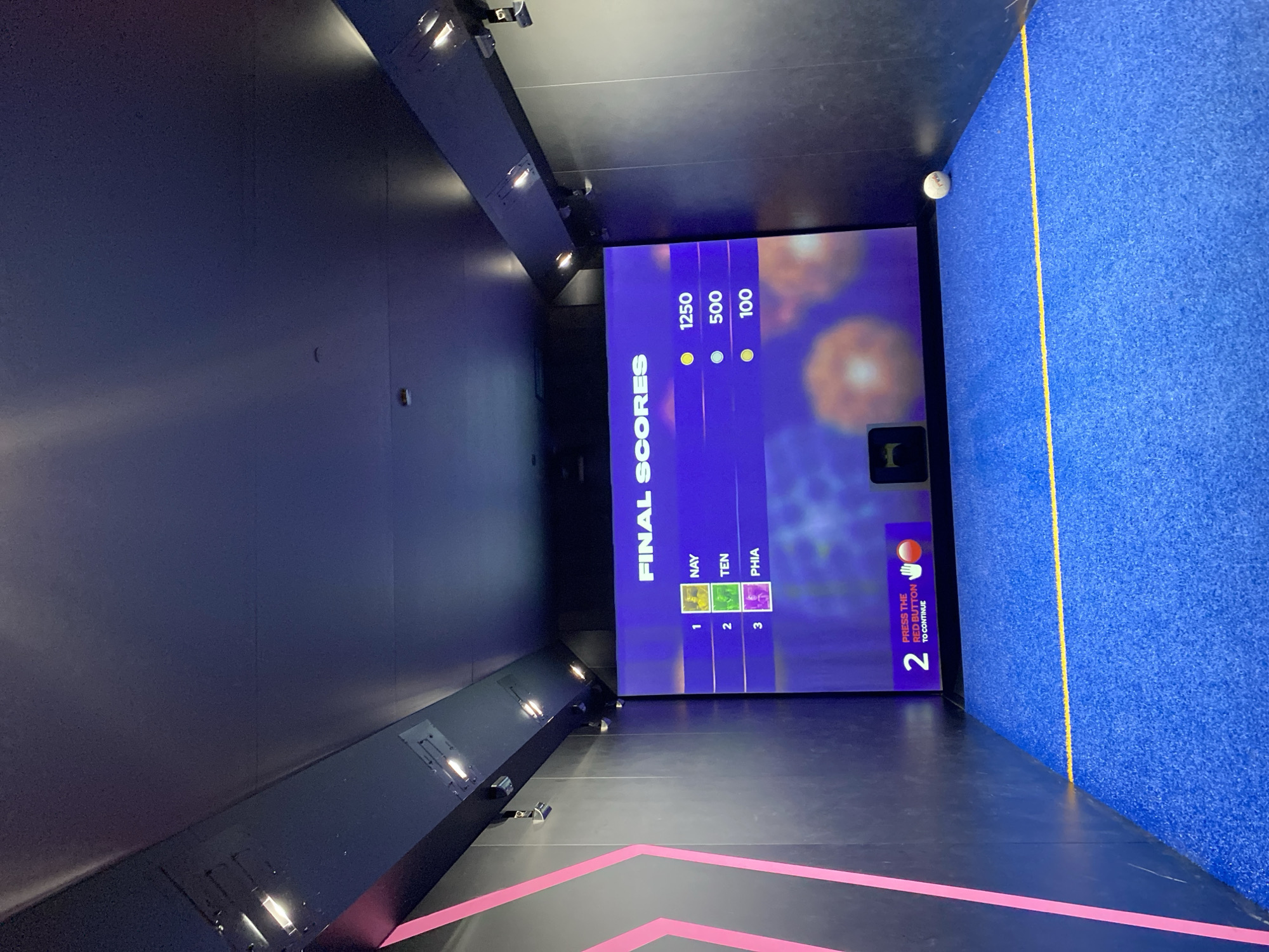 This appears to be an image of an interactive digital game, likely located in an entertainment venue in London, United Kingdom. The room has a dark, modern design with integrated lighting fixtures along the ceiling and side walls. The main feature is a large screen displaying game results, titled "FINAL SCORES" with participant names (NAY, TEN, PHIA) and their corresponding scores (1250, 500, 100). The screen also prompts the user to press the red button to continue, indicating the game is still in progress. The floor has a black surface with pink geometric patterns, and a blue textured wall stands to the right with a yellow line marking the boundary. A white ball rests on the top edge of the blue wall.