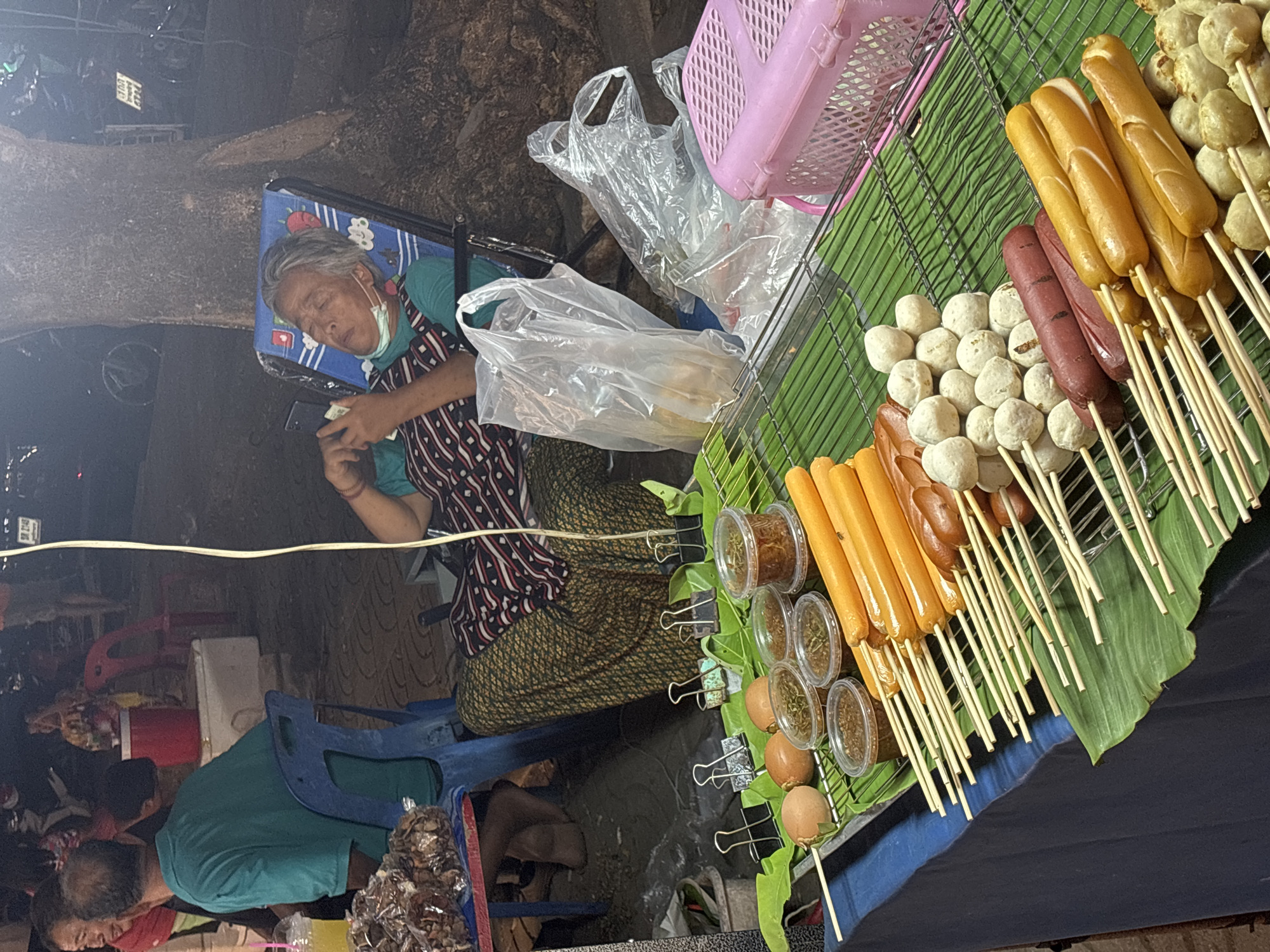 This media file captures a nighttime scene at a street food stall in what appears to be a market in Choeng Thale, Thailand.
The primary subject is an elderly woman, possibly the vendor, who is asleep or resting in a blue folding chair next to her stall. She has grey hair, a face mask pulled down, and is wearing a light blue top, a patterned apron, and a long skirt. She holds a smartphone in her left hand while her eyes are closed.
Her food stall is prominently displayed, featuring numerous skewers of sausages (both light brown and reddish-brown) and white meatballs, arranged on banana leaves over a metal grate. Several small plastic containers of sauces or seasonings, along with a few hard-boiled eggs, are also part of the display. A pink plastic basket and various clear plastic bags are visible near her.
In the background, other people can be seen, including a man in a green shirt, suggesting an active market environment. The scene is illuminated by artificial lights, indicating it is evening or night. The ground appears to be dark and unpaved. The entire image is rotated 90 degrees to the right. A small sign with "ปท 9423" is faintly visible in the upper left corner.