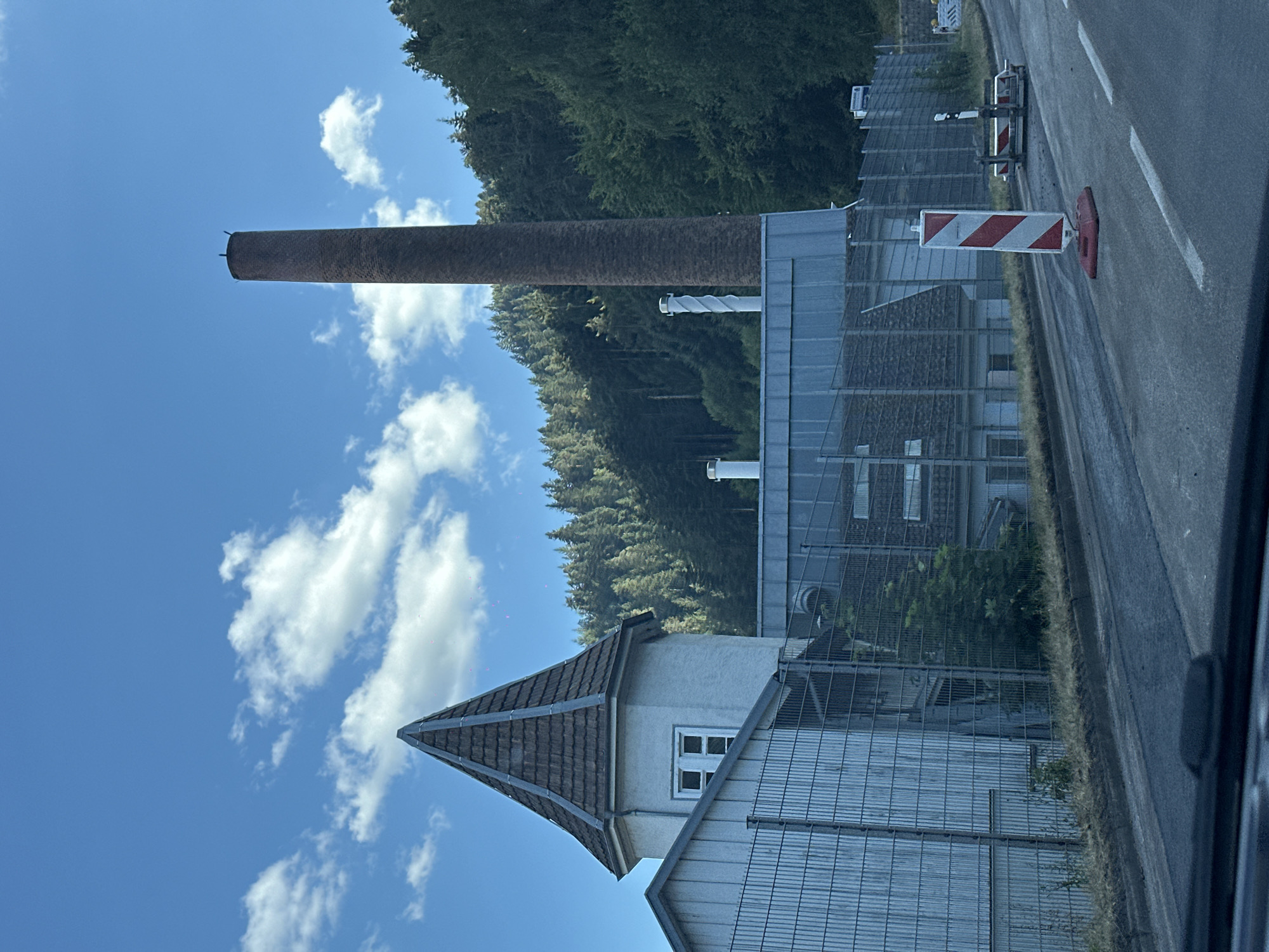 The image shows an industrial building complex with a tall, brick chimney in the background, located in Hornberg, Germany. The site is surrounded by a metal fence and situated at the base of a densely wooded hillside. A paved road with white lane markings runs in the foreground, with a temporary traffic barrier and a red and white striped pole indicating ongoing road work or a detour. The sky is blue with scattered white clouds. The architecture of the building includes a distinctive octagonal tower with a conical tiled roof and a white-paneled structure.