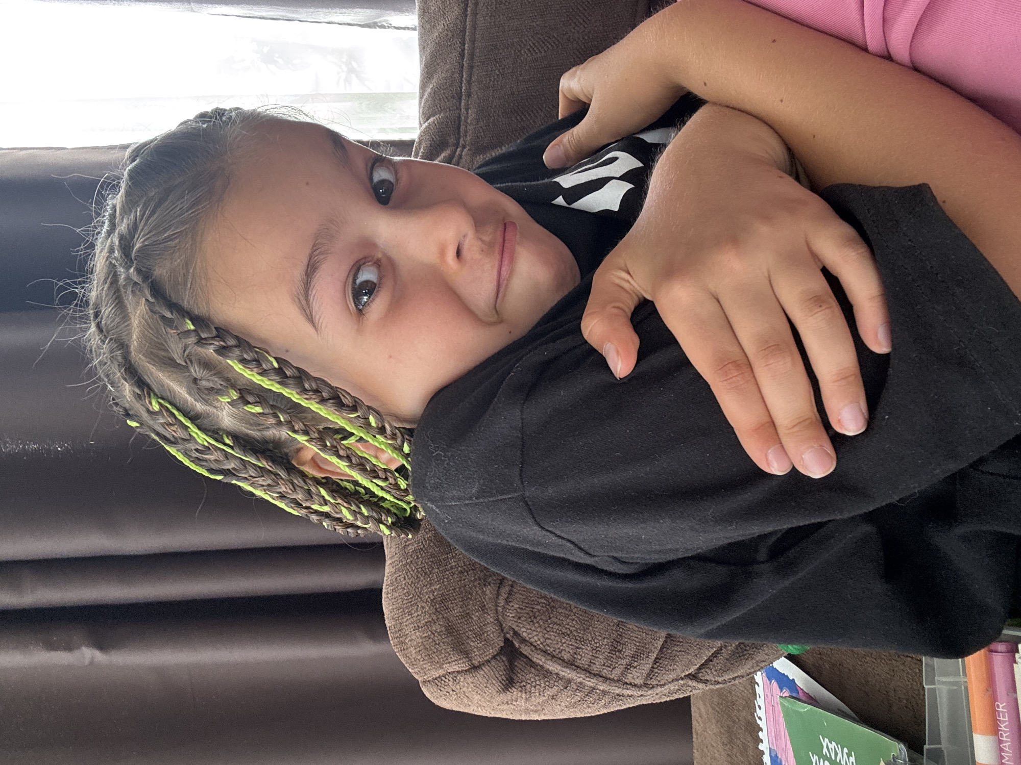 This media file shows a close-up, rotated view of a young person with braided hair, some of which is accented with bright green. They are lying down, possibly on a couch, as indicated by the textured brown fabric visible. The person is wearing a black t-shirt with a white graphic design on it. Their eyes are open and they are looking towards the camera with a playful or curious expression. Another person's arms are visible, embracing the subject, with one arm wearing a pink garment. The background appears to be indoors, near a window with natural light filtering through, suggesting it is daytime. In the lower right corner, the corner of a clear plastic container holding art supplies like markers can be seen. Some visible text on the art supplies includes "РУКАХ" (which translates to "hands" in Ukrainian) and "MARKER". The overall mood is relaxed and intimate. The context of "Uyutnoye, Ukraine" suggests the location where this image was taken.