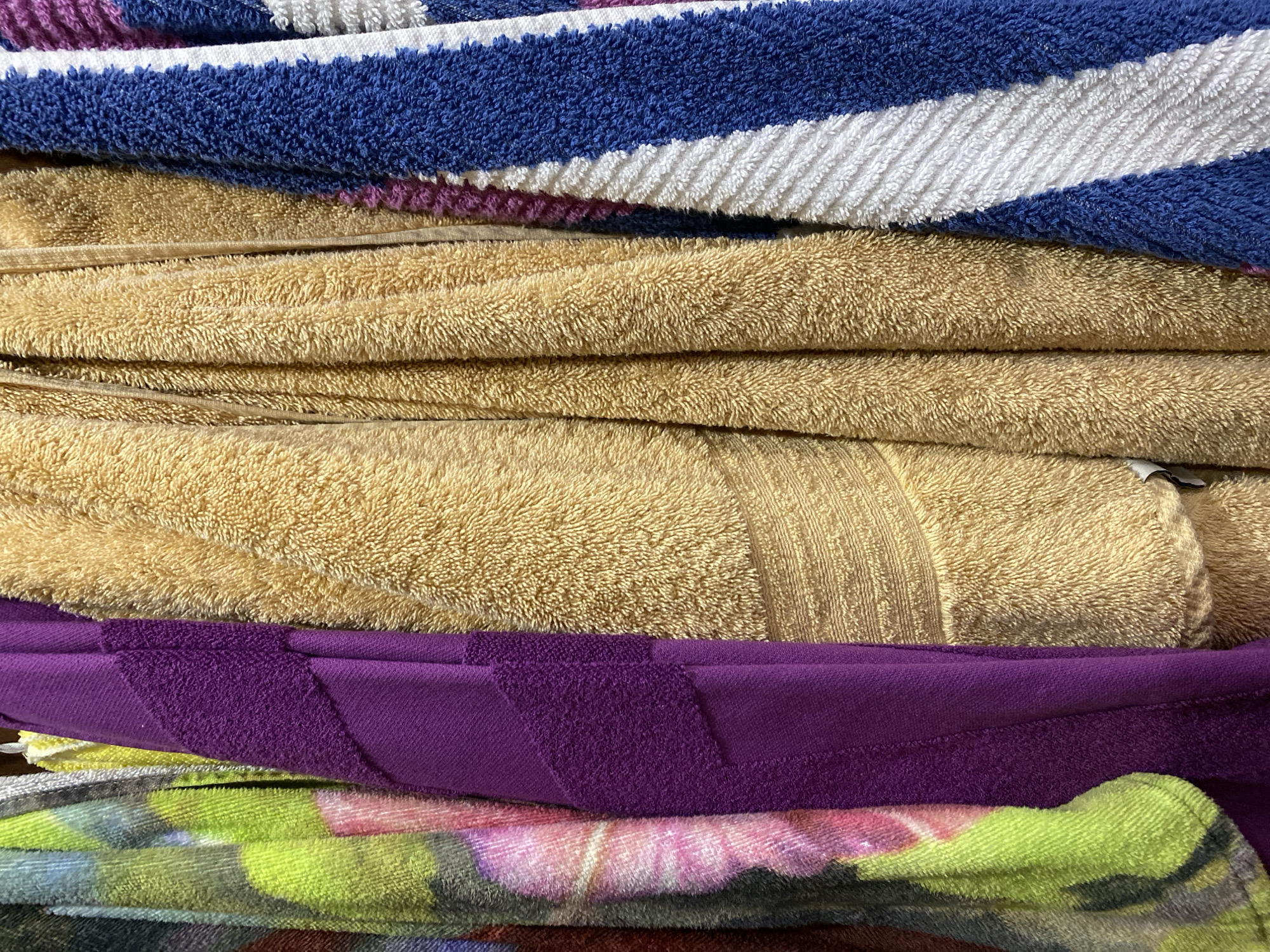 This is a close-up, overhead shot of a stack of folded towels. The towels are layered on top of each other, creating a textured surface. The topmost towel has diagonal stripes of blue and white, with thinner stripes of purple and white. Beneath that are two solid tan towels, one with a ribbed texture. The next towel is a deep purple with a textured pattern. Below that is a towel with a floral pattern in shades of green, pink, and yellow. The bottom of the stack is partially visible and appears to be a dark, patterned towel. The lighting suggests it is daytime, and the setting is likely domestic, possibly a linen closet or laundry room. There is no text visible in the image.