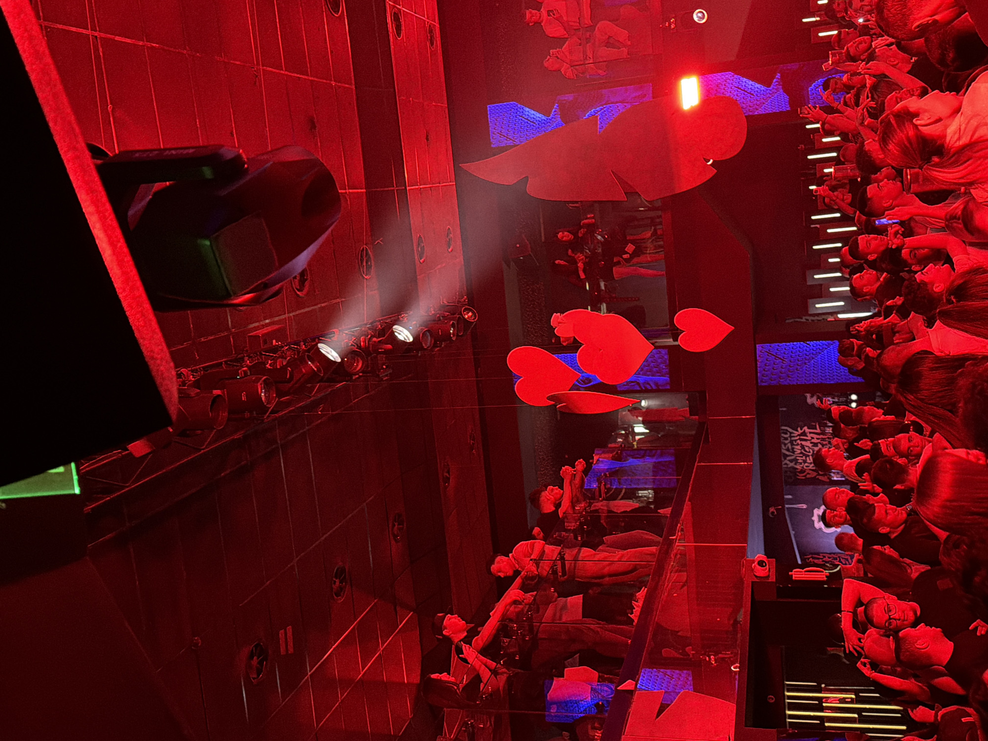 The image captures a vibrant indoor event, likely a concert, club night, or performance, illuminated predominantly by intense red lighting, with accents of blue. The scene is bustling with a large crowd of people, many of whom are looking towards an unseen stage or focal point, with numerous individuals holding up smartphones, possibly recording or taking photos.
On the left side of the frame, a segment of the venue's structure is visible, featuring dark walls with circular vents or speakers, and mounted stage lights emitting bright beams that are colored red by the ambient light. A large, dark projector or moving head light fixture is prominent in the upper left. Several decorative red heart-shaped cutouts are suspended in the air.
Towards the right, more attendees are visible, some sitting at tables with drinks, while others stand. The strong red lighting casts a dramatic glow on their faces and surroundings. Reflections of the crowd and lighting are visible on what appears to be a polished floor or mirrored surface. In the background, on a dark wall, some graffiti-style text is partially discernible, with "REGAL" being somewhat legible, alongside other faint letters.
The atmosphere appears lively and engaging, consistent with a nighttime entertainment event in a modern venue. The location context given is Blagoevgrad, Bulgaria.