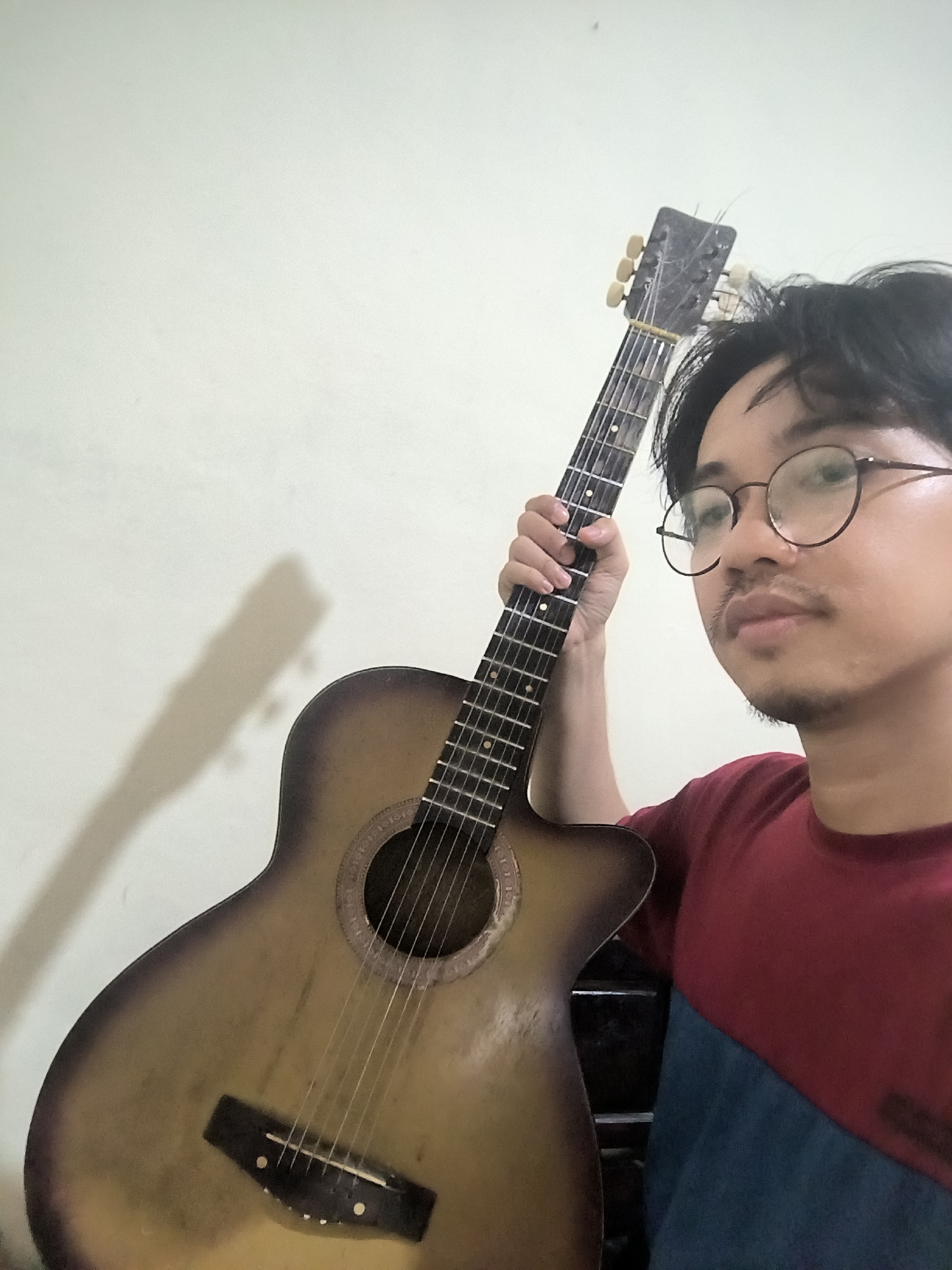 A man with glasses and a mustache holds an acoustic guitar. He is wearing a red and blue shirt. The guitar is positioned vertically, with the man's left hand on the fretboard. The background is a plain, light-colored wall with a shadow of the guitar and the man cast upon it. No specific location in Bumi Mulyo, Indonesia, can be identified from the image.