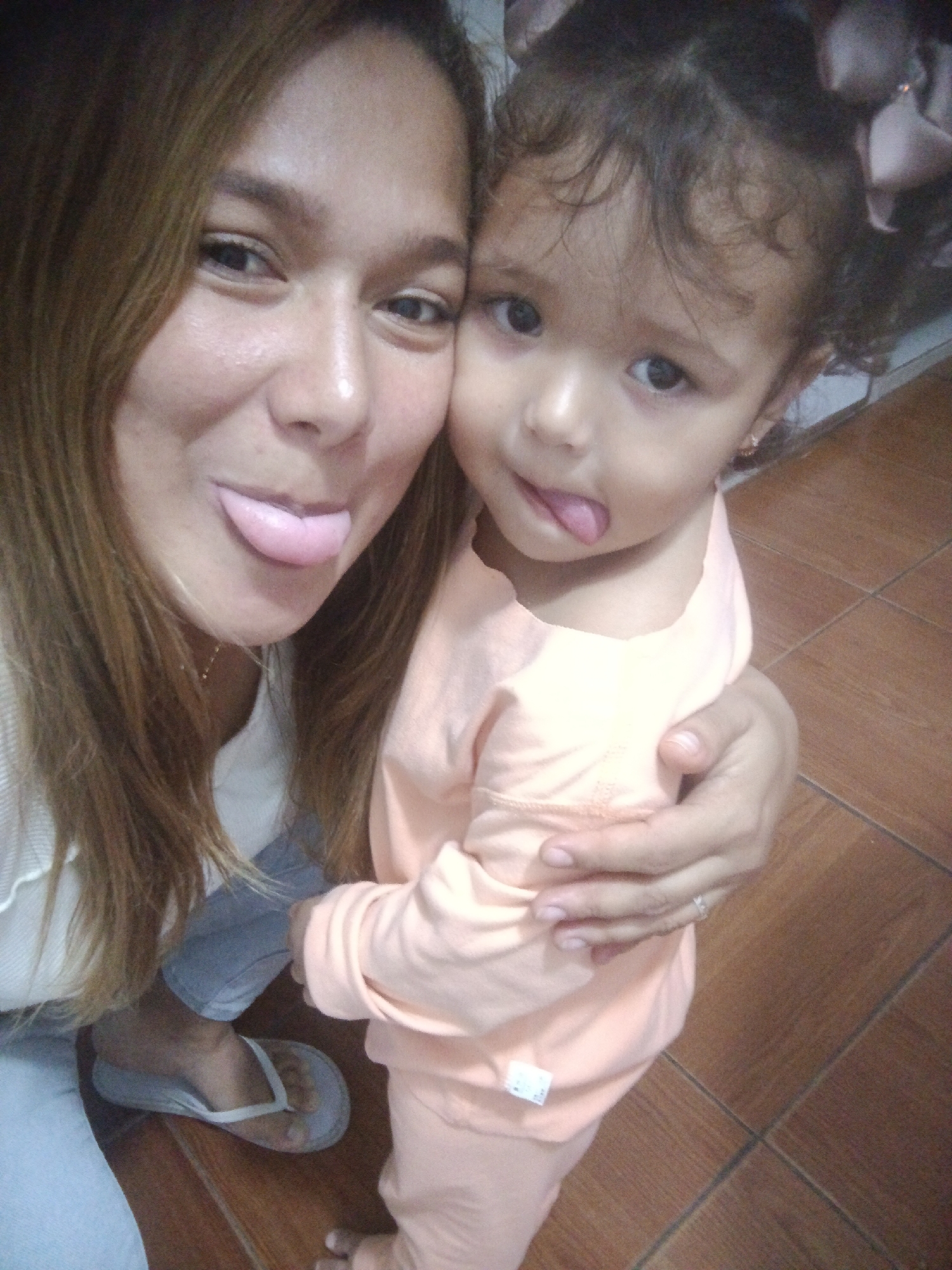 A woman and a young child are taking a selfie. The woman has long brown hair and is wearing a white shirt. The child has curly brown hair and is wearing a peach-colored shirt. Both are sticking out their tongues. They are indoors, with a tiled floor visible. The image is likely taken in a residential setting in Caracas, Venezuela, given the context of the prompt.