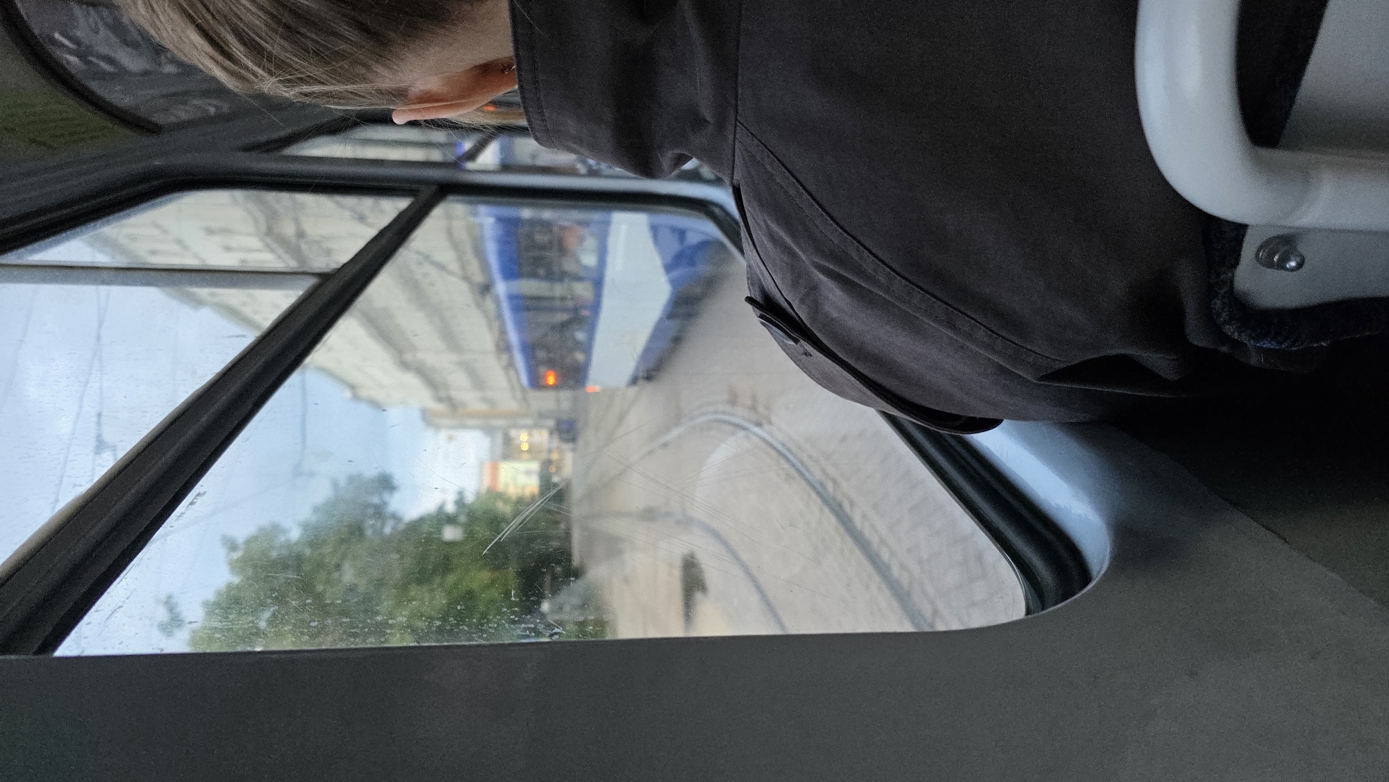 The image, taken from inside a tram in Riga, Latvia, shows a view through a wet window. A person wearing a dark jacket is seated on the right, facing forward. Outside the tram, a blue and white tram with illuminated windows is visible in the distance, traveling along tram tracks on a street lined with buildings. Trees are also present on the left side of the frame. The sky appears overcast.