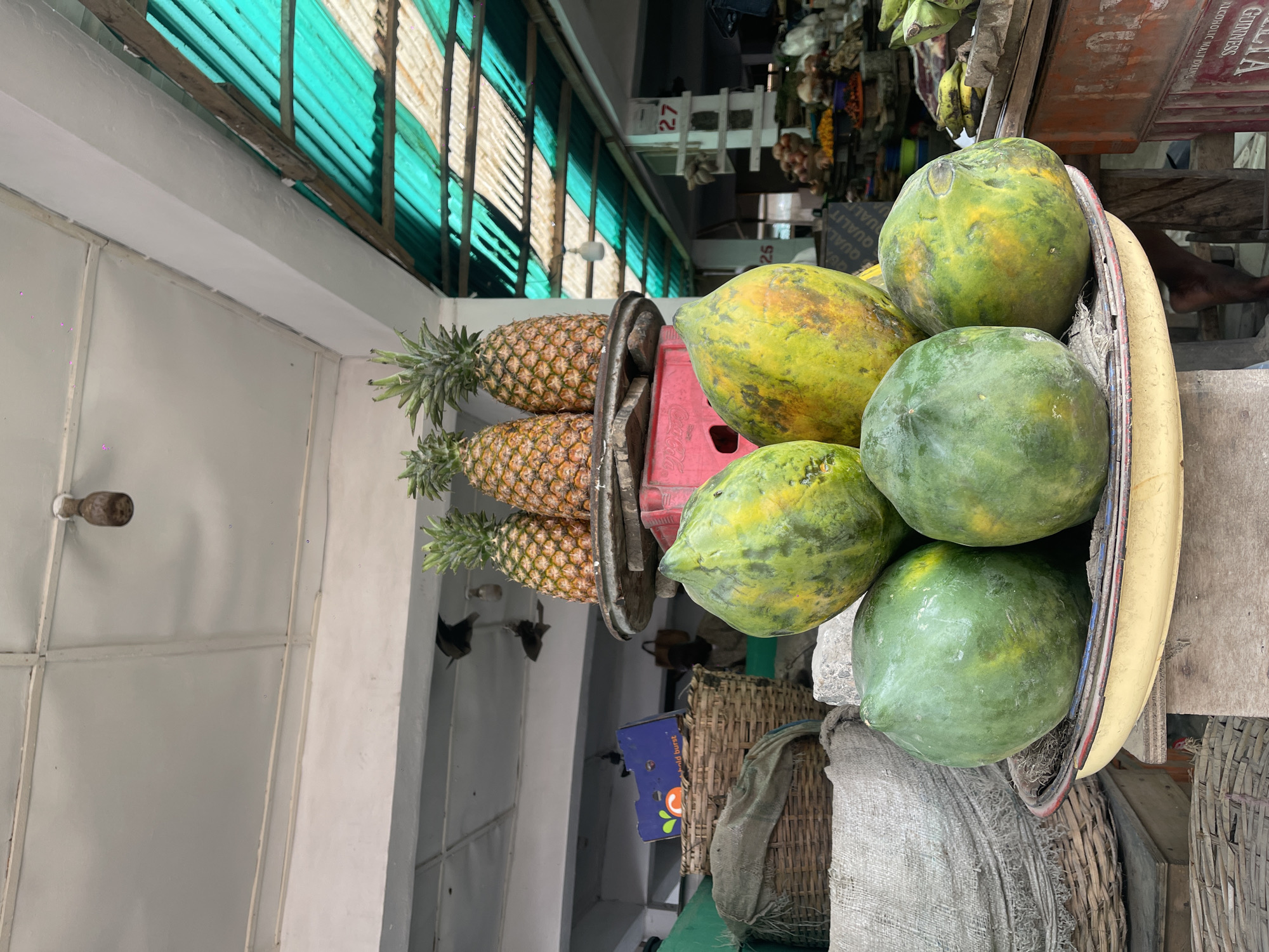 This image captures a vibrant, sunlit produce stall within what appears to be an indoor or covered market. The scene is dominated by a display of fresh fruit.
In the foreground, a generous pile of ripe and unripe papayas, predominantly green with patches of yellow, rests in a yellow basin or tray. Some of these papayas are supported by a red plastic crate, partially visible. To the left of the papayas, three pineapples with their spiky green crowns are stacked horizontally on a stand, showcasing their golden-brown, patterned skins. Below the fruit display, woven baskets and rough burlap sacks are visible, suggesting storage or additional goods.
The background reveals more of the market environment. A distinct corrugated metal roof, with alternating green and white translucent panels and wooden support beams, extends across the top of the image, allowing natural light to filter in. Further back, various market goods are blurred, and a white stall structure with the red number "27" is noticeable. Other faint objects, including what appears to be hanging produce like onions, add to the market atmosphere. A bare foot is partially visible on the far right, near the fruit display, indicating the presence of a person, likely a vendor or customer. Visible text includes "27" on the stall, a partial "GUINNESS" and "ALCOHOLIC MALT DRINK" on a sign, and "QUALITY QUALIT" on another label.
The bright lighting suggests it is daytime. The overall impression is one of a busy, functional market stall in a tropical or subtropical region, offering fresh local produce.