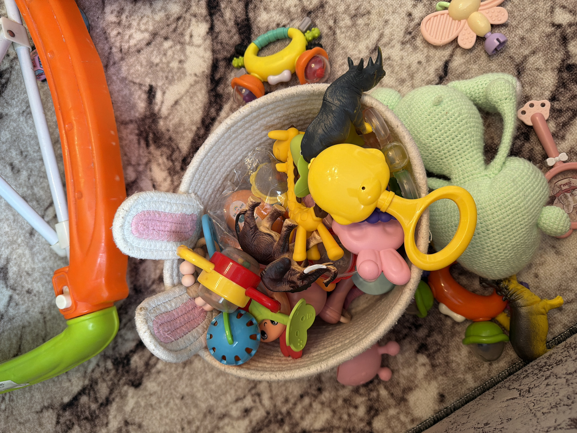 In Sovkhoza "Razdol'e", Russia, an overhead shot captures a collection of baby toys scattered across a patterned carpet. A woven basket, decorated with plush bunny ears, is filled with various items including rattles, animal figures (a dinosaur, a deer, and an elephant), and colorful plastic toys. Outside the basket, more toys are visible, such as a large green crocheted plush toy, a pink butterfly-shaped teether, and a red and orange plastic toy walker. The lighting is natural, suggesting daytime.