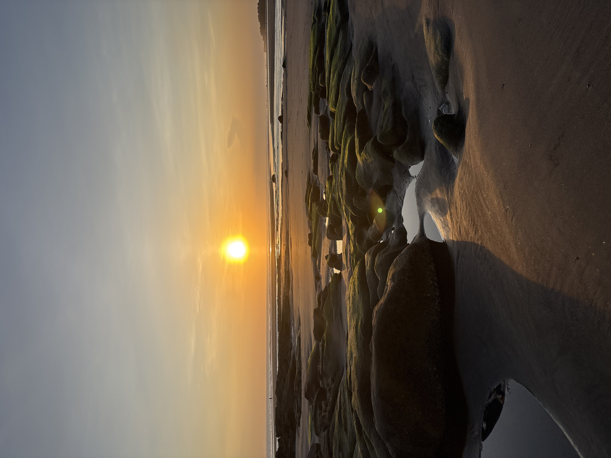 The image shows a sunset view at a beach in Kerambitan, Indonesia. The sun, a bright yellow orb, is positioned low in the sky, casting a golden light across the scene. The sky transitions from a pale yellow near the horizon to a light gray further up. The beach features dark rocks covered in green moss in the foreground. Small pools of water reflect the sunlight. The wet sand glistens. In the distance, a sandy beach stretches along the shoreline, with the ocean faintly visible. A single figure is seen on the beach. The scene is mostly still and serene, depicting a quiet moment at sunset.