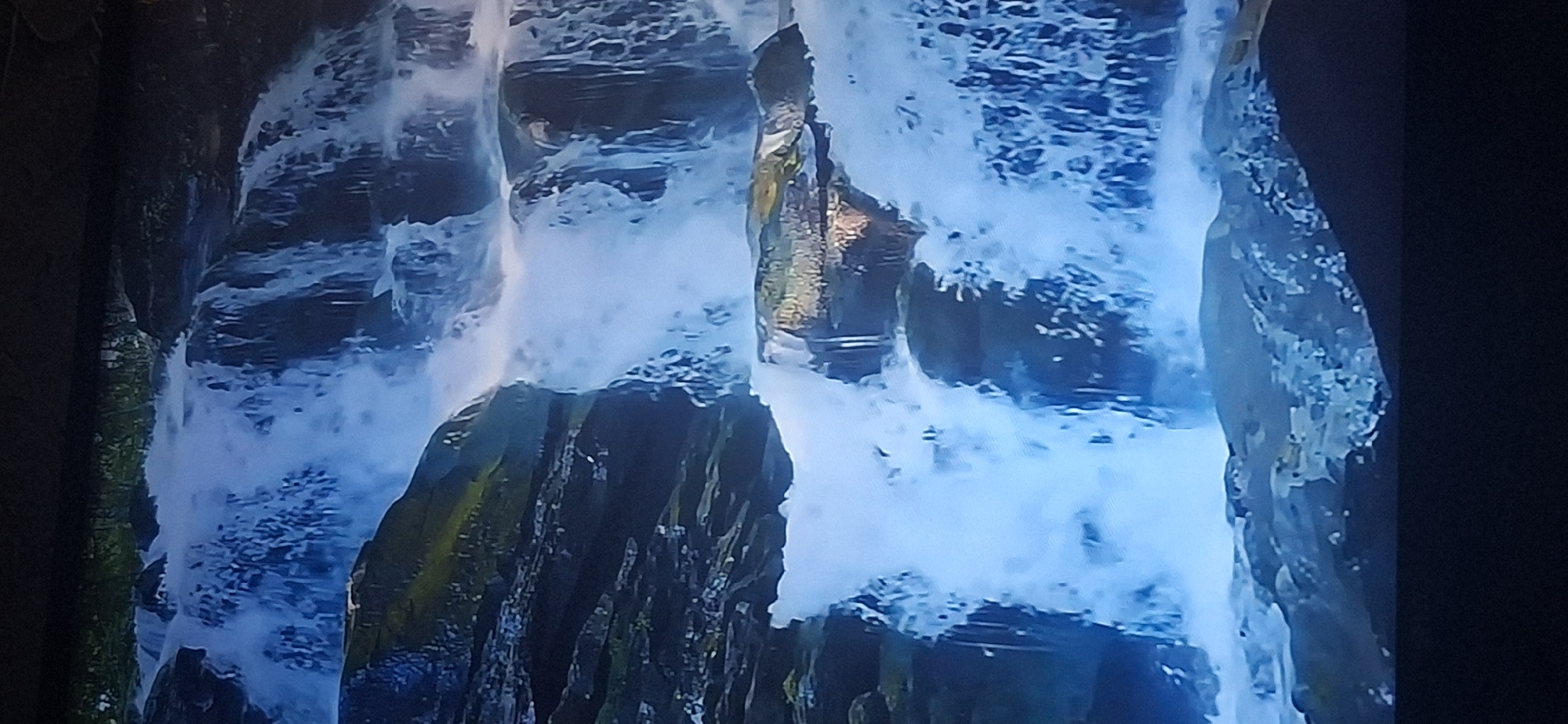 The image shows a multi-tiered waterfall cascading over dark, rocky ledges covered in moss and vegetation. The water is a bright white and appears frothy and turbulent as it tumbles down. The surrounding environment is lush and green, with hints of sunlight filtering through. There are no people or man-made structures visible in the image. This image does not depict a location in the city of Bila Tserkva, Ukraine.