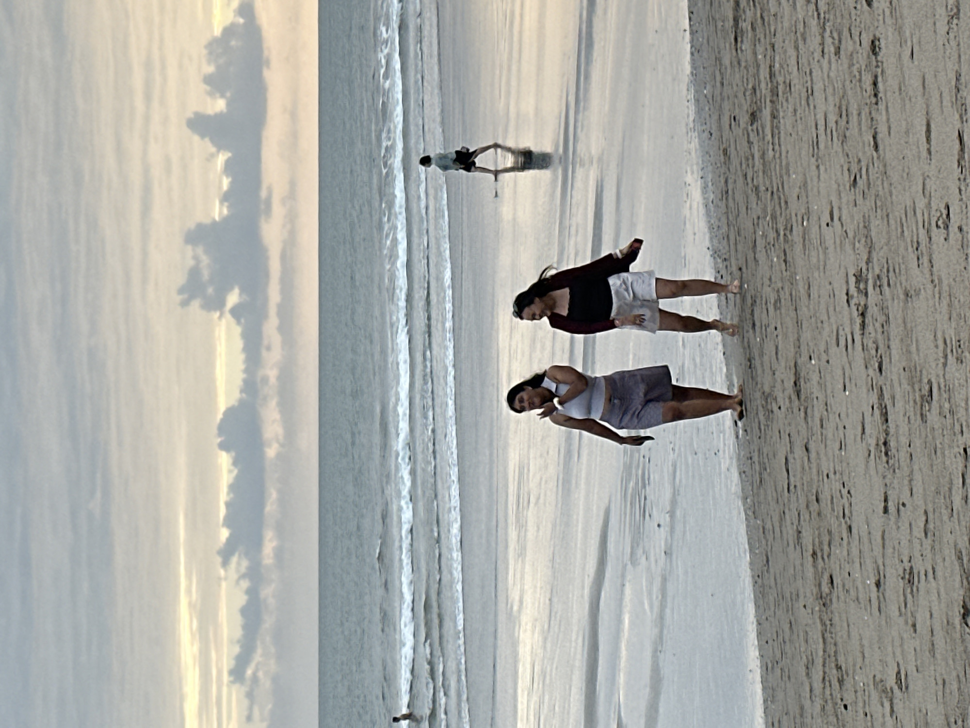 Two women are walking on a beach in North Kuta, Indonesia, near the ocean. The woman on the left is wearing a light-colored top and a patterned skirt. The woman on the right is wearing a black top, a maroon cardigan, and white shorts. They are walking side-by-side and appear to be talking and gesturing. The beach is wet and reflects the sky, indicating it has recently rained or the tide is high. The ocean has gentle waves, and the sky is cloudy with hints of the sun breaking through. In the background, there are other people on the beach and in the water. The overall scene depicts a casual moment at the beach.