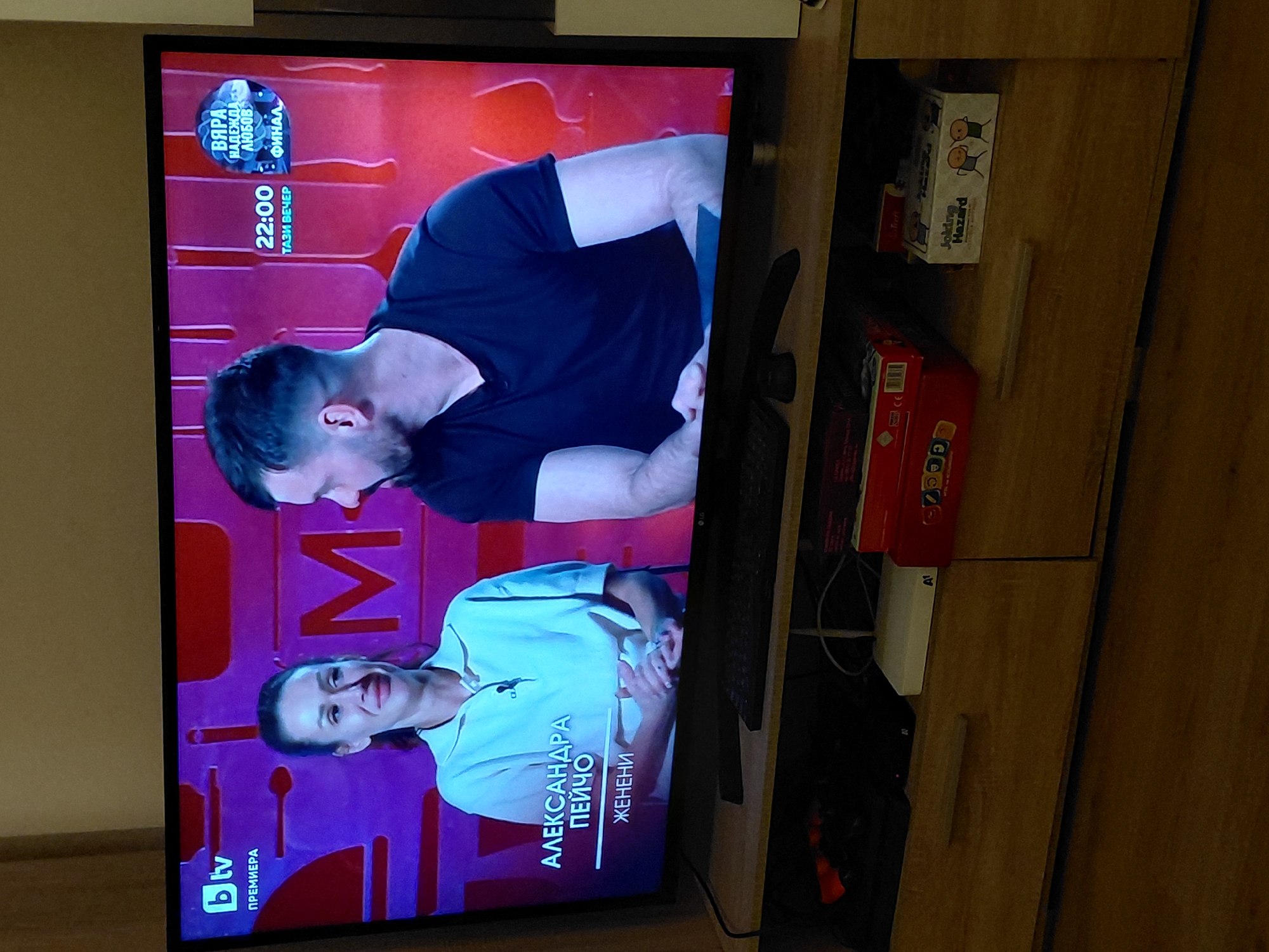 A television displays an episode of the Bulgarian reality television show "Married at First Sight" (Женени от Първ поглед), featuring the names "Александра Пейчо" and showing the logo of the BTV television channel, indicating it is airing in Sofia, Bulgaria. There are game boxes and a media box next to the television on a wooden entertainment center.