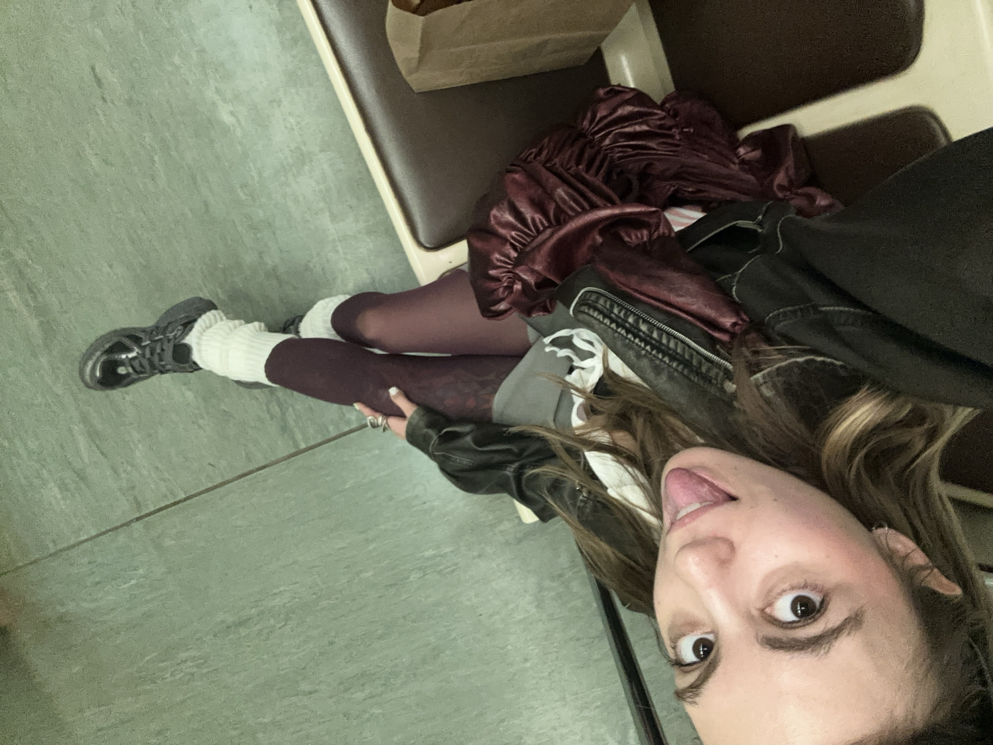 This media file shows a young woman, likely in her late teens or early twenties, posing for a selfie. She is seated on a bench that appears to be in a public transport vehicle, possibly a bus or a train, given the upholstery and the flooring. The setting suggests an indoor environment with artificial lighting, and there are no clear indications of the time of day or weather conditions.
The woman is wearing a dark, possibly black, leather jacket over a gray top. She has on burgundy-colored tights with a patterned texture and white, ribbed knee-high socks. Her footwear consists of dark-colored sneakers. A ruffled, shiny, burgundy skirt or dress is draped over her legs.
Her face is turned towards the camera, and she is sticking her tongue out, with her eyes wide open. She has long, light brown hair that falls around her face. She is wearing rings on her fingers and earrings. The overall mood conveyed by her expression is playful and informal.
The background shows a segment of the bench, some brown upholstery, and a crumpled paper bag. The flooring is a light green, mottled material. There is no visible text in the image. The context provided mentions "Moscow, Russia," which might imply the setting is a public transport system within that city.