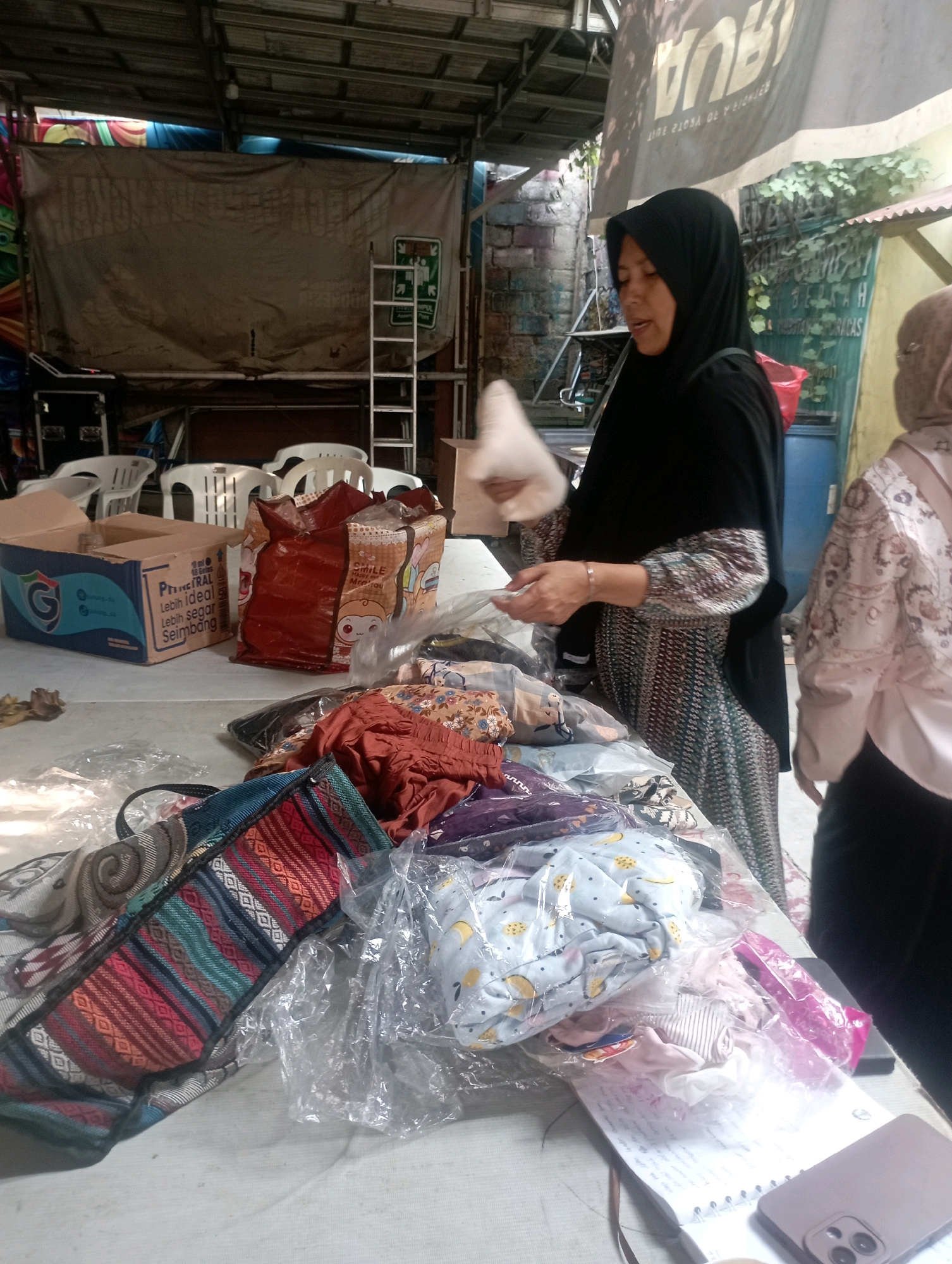 A woman wearing a black hijab and a patterned dress stands at a table laden with folded clothes and a patterned bag. She is looking down and appears to be handling a white item. Another woman, partially visible on the right, wears a patterned hijab and light-colored top. The table is covered with plastic-wrapped clothing, some of which is colorful and patterned. A red bag and a cardboard box are also present on the table. In the background, a ladder leans against a large tarp, and several white plastic chairs are stacked. The scene appears to be outdoors under a metal roof structure, suggesting a market or outdoor vendor setting in East Jakarta, Indonesia.