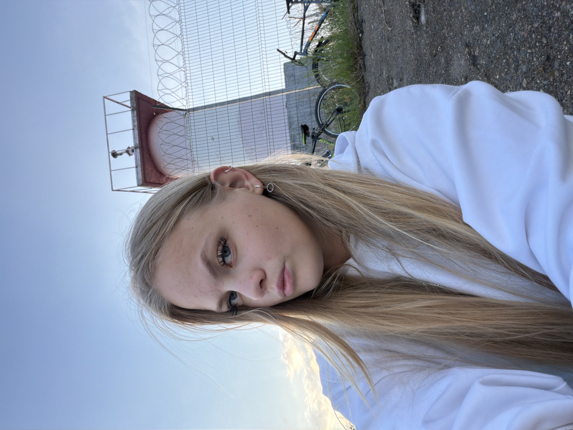 In a selfie taken outdoors, a young blonde woman with blue eyes and light skin, wearing a white hoodie, faces the camera. Her hair is windswept, and she has small hoop earrings in her left ear. Behind her, a lighthouse with a pink base and a metallic top stands against a clear blue sky with wispy clouds. A barbed wire fence runs horizontally behind the lighthouse, and two bicycles are parked on the grassy ground. The ground in the foreground is paved with rough asphalt. The scene appears to be located near the Kama River in Chaikovsky, Russia, given the distinct lighthouse structure often seen in the city.