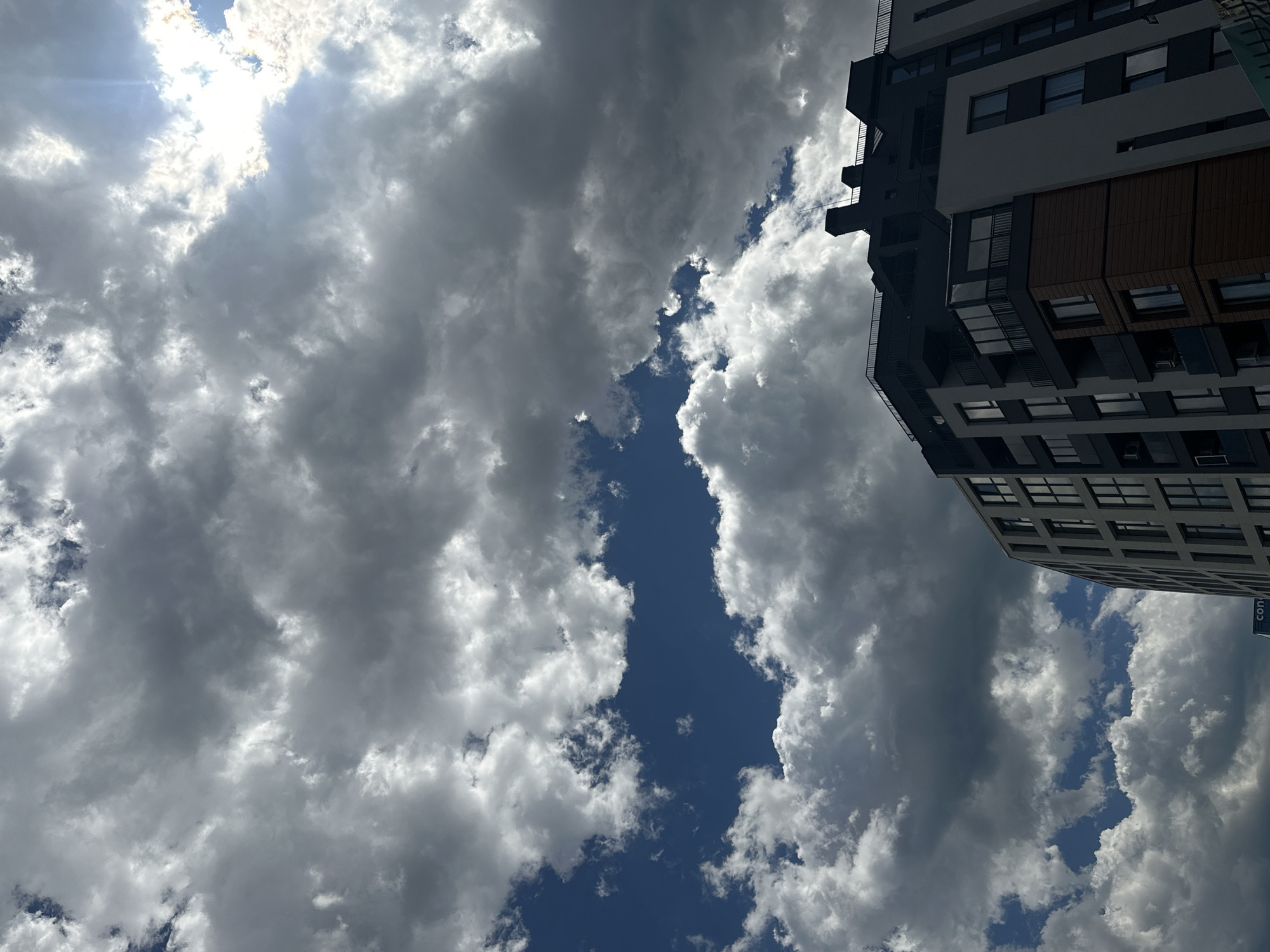 A low-angle shot captures a modern, multi-story building against a dynamic sky filled with dramatic clouds. The building, partially silhouetted, features a dark upper section with a railing and chimneys, contrasting with lighter-colored sections below. The sky is a mix of bright blue patches and dense, dark gray clouds, with the sun peeking through the upper right, creating highlights and shadows. The lower right corner of the image shows the edge of another building and a fence. The scene appears to be an urban environment, possibly in Sofia, Bulgaria, given the architectural style and the presence of modern construction.