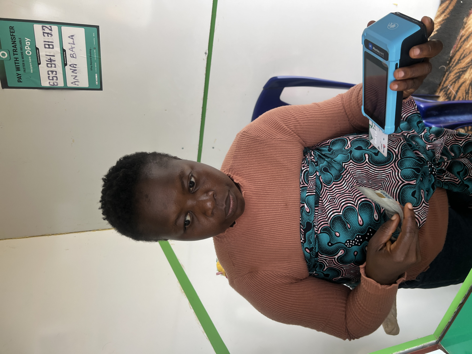 A medium shot captures a woman of African descent in Gura Top, Nigeria, focused on a transaction. She is wearing a rust-colored, ribbed long-sleeved shirt and a vibrant teal, pink, and black patterned wrap around her waist. Her left hand holds a card inserted into a blue point-of-sale (POS) terminal, which she is operating with her right hand. The POS terminal is black and blue, with a screen displaying a logo that reads "Mon Evelyn." In the background, a white wall with a green stripe runs horizontally. A blue chair is visible behind the woman, and a sign on the wall above her right shoulder displays "PAY WITH TRANSFER" and "O Pay," along with an account number and name. The lighting is bright, suggesting an indoor setting.