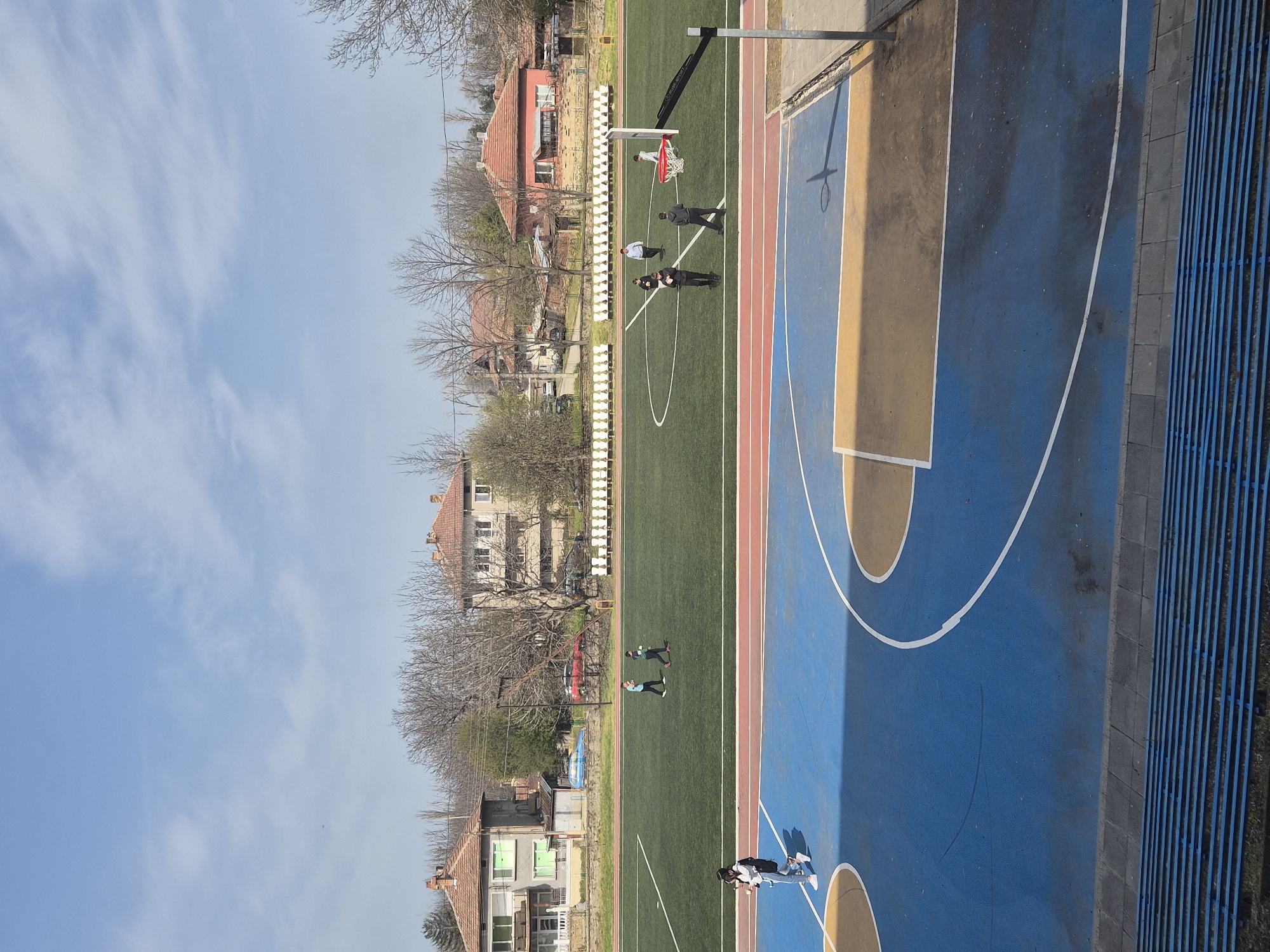 The image depicts an outdoor sports complex in Pavlikeni, Bulgaria, featuring a basketball court with blue and tan painted sections and white lines. Adjacent to the basketball court is a green artificial turf field marked with red lines. White stadium-style seating lines one side of the field. Several people are on the field; some appear to be standing near a basketball hoop, while others are walking. Residential buildings with red tile roofs and bare trees are visible in the background. The sky is blue with some scattered clouds.