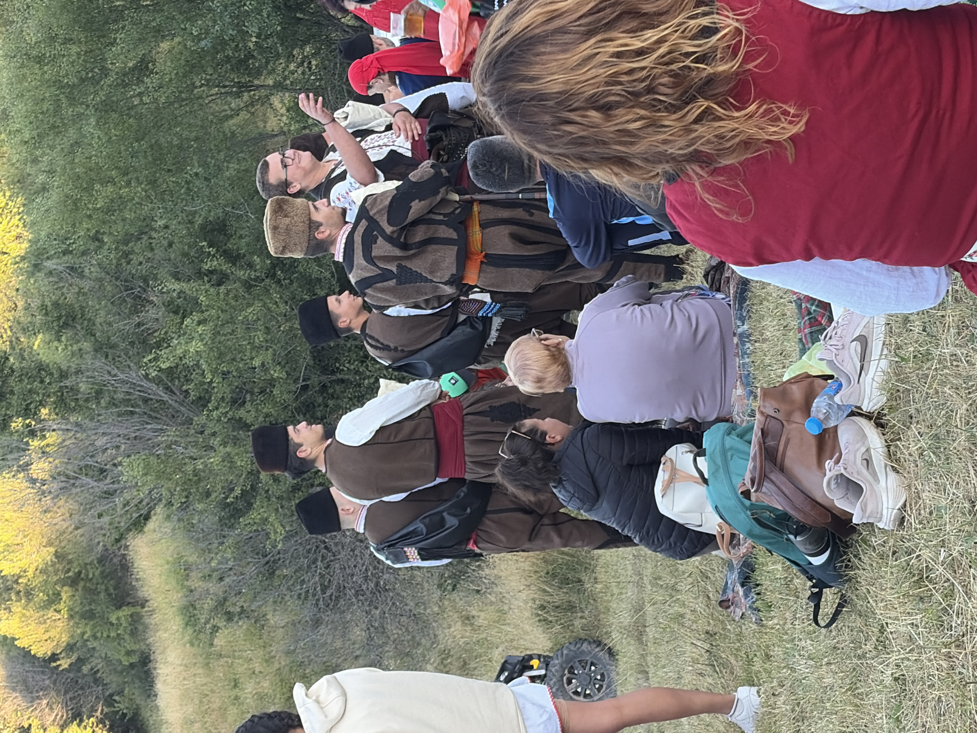 The image depicts an outdoor gathering in the city of Magardjitsa, Bulgaria, with a group of people dressed in traditional attire. The scene appears to be set in a grassy, hilly area with trees in the background, suggesting a rural or park-like setting.
In the foreground, several individuals are seated or standing. One person, viewed from behind, has long, wavy blonde hair and wears a red vest over a white shirt. To their left, a person in a purple top is seated, facing away from the camera. Beside them, a person in a dark jacket is also seated. A collection of bags and shoes are scattered on the ground near these seated individuals.
A group of men in traditional Bulgarian folk costumes are a prominent feature in the mid-ground. They wear dark vests over white shirts, with colorful sashes around their waists and traditional hats. Some of these men are engaged in conversation or observing something to their right. One man in a brown vest and patterned tunic is holding a green object. Another man, wearing a fur-trimmed hat, is gesturing with his right hand.
Further to the left, a person in a light-colored hoodie and white shorts is standing near a small all-terrain vehicle. The overall atmosphere suggests a cultural event or festival. The lighting indicates it is either late afternoon or early morning, with sunlight filtering through the trees.