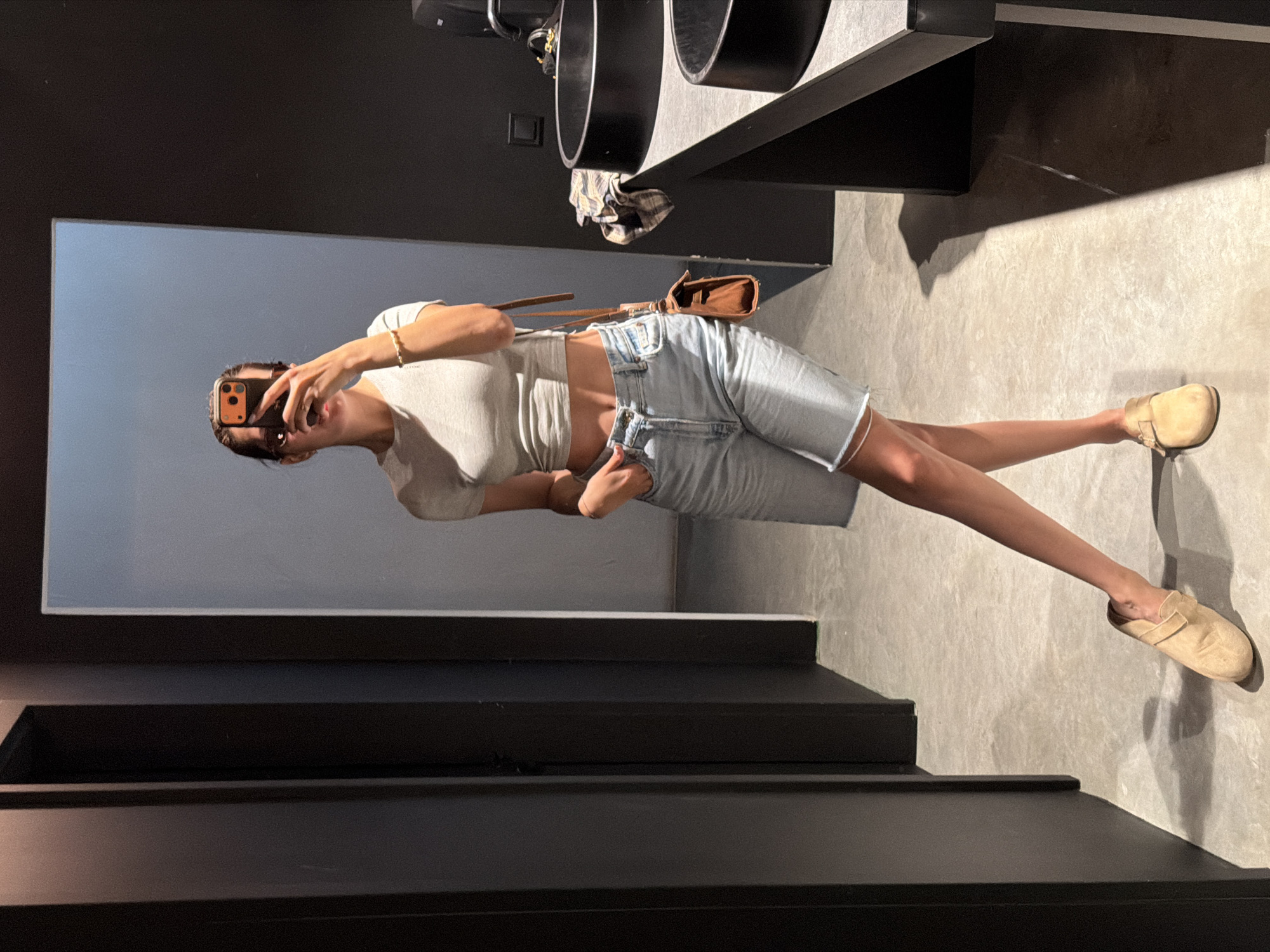 A young woman with dark hair pulled back is taking a mirror selfie in a modern bathroom. She is wearing a light-colored crop top and light-wash denim shorts. A small brown crossbody bag hangs from her right shoulder. She has her left hand in the pocket of her shorts and is holding a phone in her right hand, obscuring her face. She is wearing tan slip-on shoes. The bathroom has dark walls, grey tiled floors, and a dark countertop with black sinks.