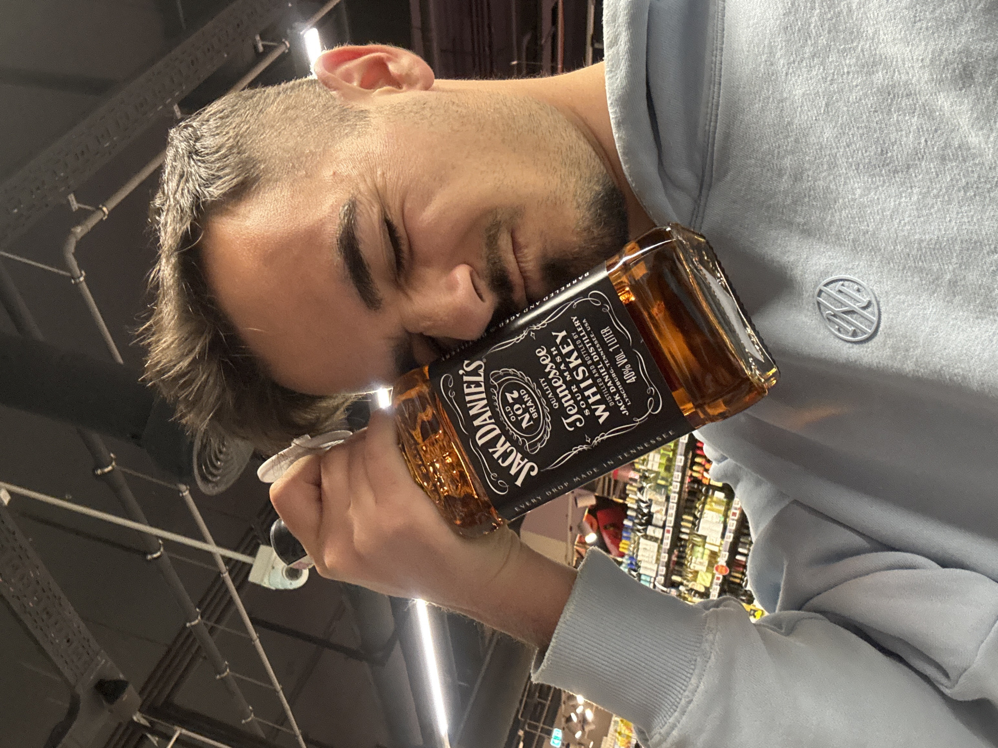 This image, taken in Sofia, Bulgaria, captures a young man from a low-angle perspective, with his head tilted sideways and eyes closed, appearing to lean his face against a bottle of Jack Daniel's Old No. 7 Tennessee Whiskey. He is wearing a light blue hoodie that features a subtle white circular logo embroidered on the left chest. His right hand is seen holding the neck of the 1-liter whiskey bottle.
The bottle's label is prominently displayed, showing text such as "JACK DANIEL'S OLD NO. 7 BRAND QUALITY Tennessee SOUR MASH WHISKEY" and "DISTILLED AND BOTTLED BY JACK DANIEL DISTILLERY LYNCHBURG, TENNESSEE, USA 40% VOL. 1 LITER EVERY DROP MADE IN TENNESSEE."
The setting appears to be an indoor commercial environment, likely a supermarket or liquor store, with brightly lit shelves filled with other bottles visible in the blurred background. The ceiling features industrial pipes and structures, typical of such a space. The man has short, dark hair with faded sides and stubble on his face. His pose, with eyes closed and face against the bottle, suggests either a humorous exaggeration of affection for the whiskey or perhaps simulated exhaustion.