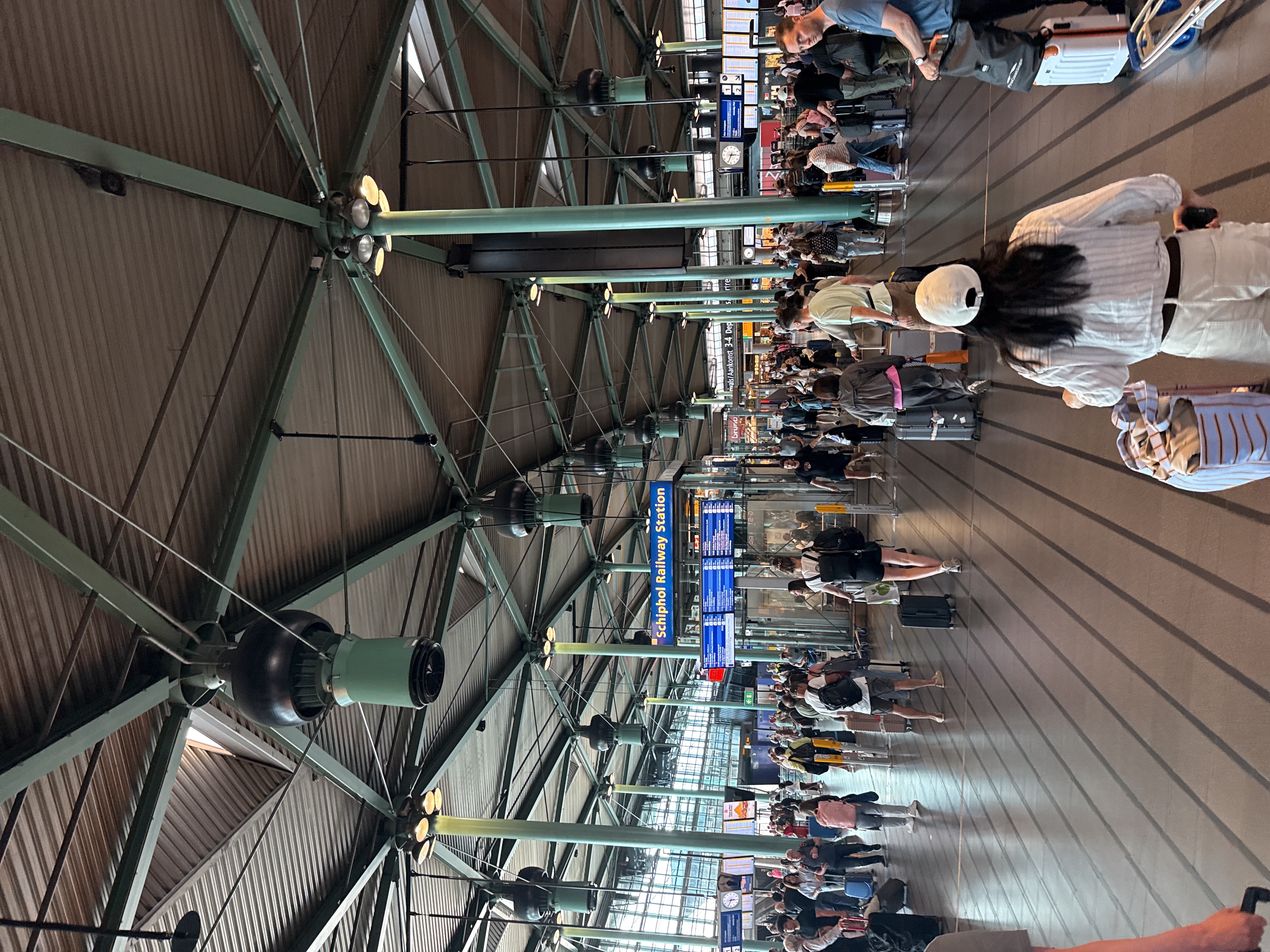 The image depicts a bustling scene inside the Schiphol Railway Station in the city of Schiphol, Netherlands. The station is populated with numerous people, many pulling luggage, suggesting they are travelers. The focus is on the interior architecture, characterized by a high, green-painted, steel-framed ceiling with integrated lighting and dark ventilation or speaker systems. Signage indicating "Schiphol Railway Station" is visible, along with train departure and arrival information displays. The floor has parallel lines that run into the distance, contributing to the sense of movement and direction in the station. The general impression is one of a busy transportation hub.
