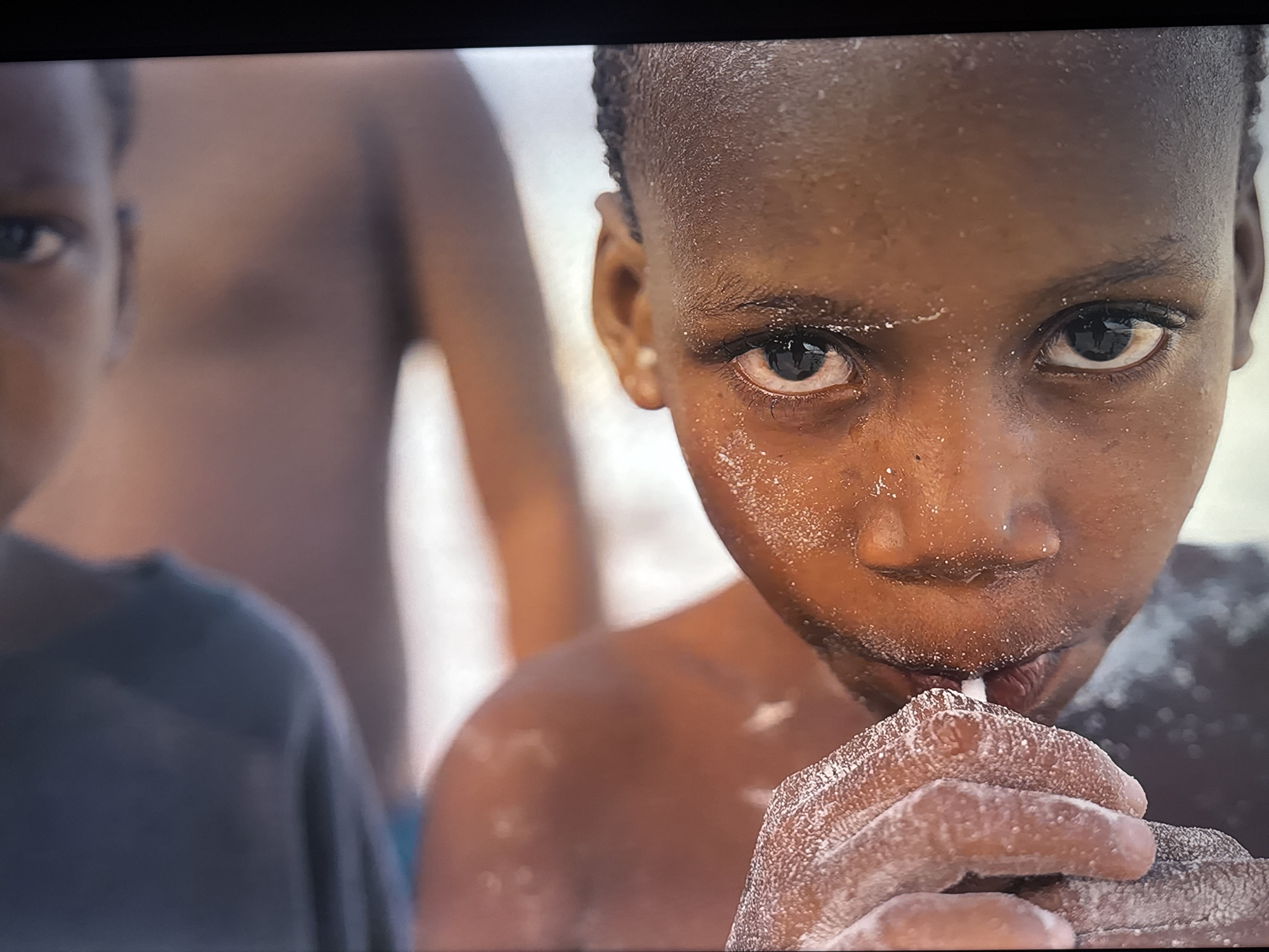 The image shows a close-up of a young Black child's face, with another child out of focus in the background on the left. The child in the foreground has wet skin, possibly from sweat or water, and is holding a white straw to their mouth, appearing to suck on something from it. Their hands are also covered in a white substance, likely powder or sugar. The background is blurred, but suggests an outdoor setting with bright light. The overall impression is one of a child enjoying a sweet treat in a warm environment.