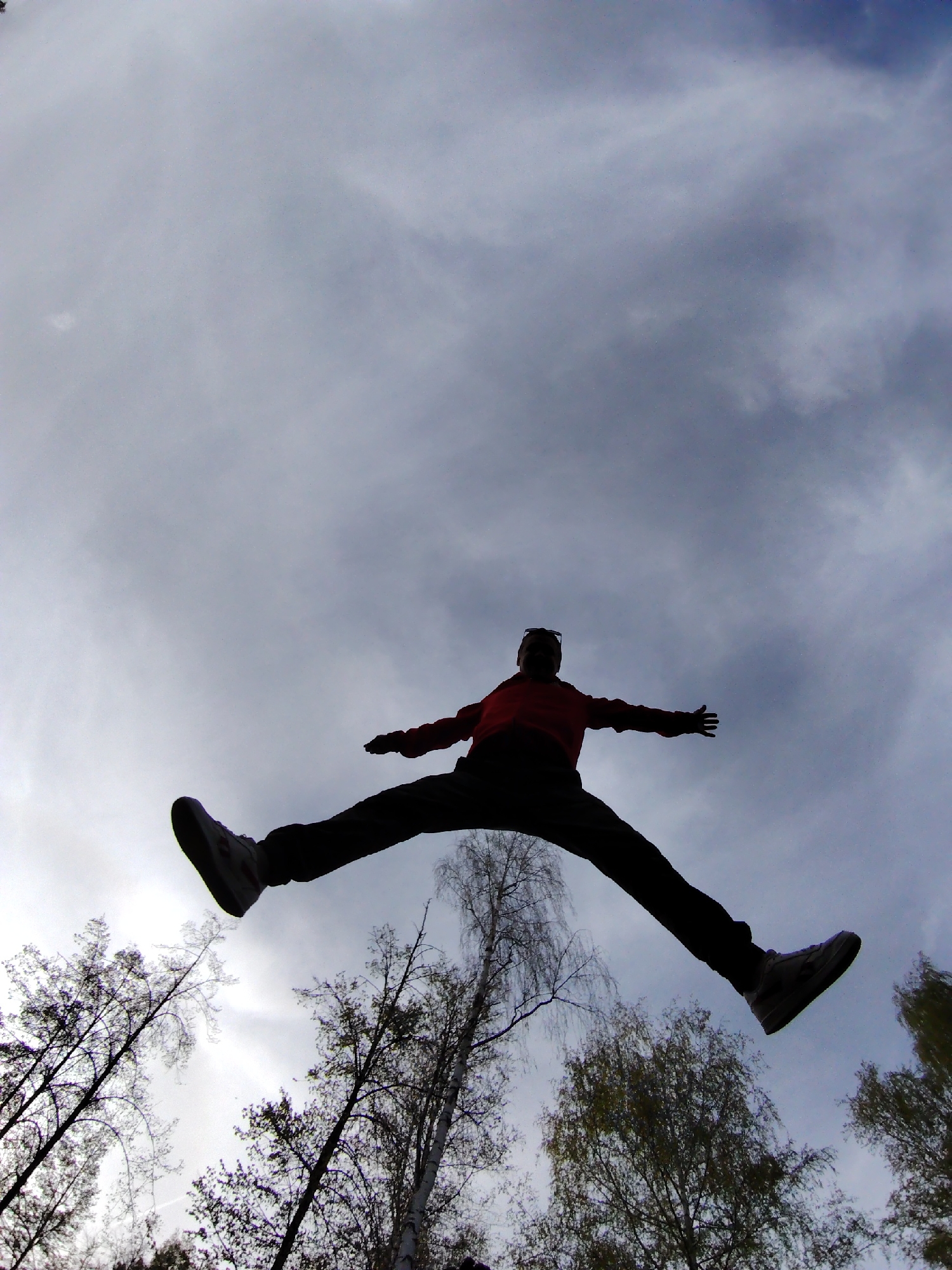 The photograph captures a person mid-air in a wide jumping stance against a backdrop of a cloudy sky and trees. The person, wearing dark pants, a red shirt, and sneakers, appears silhouetted due to the lighting conditions. The trees, with mostly bare branches, are visible from a low angle, giving the impression of looking up into the sky. The photograph was taken in Chaikovsky, Russia.