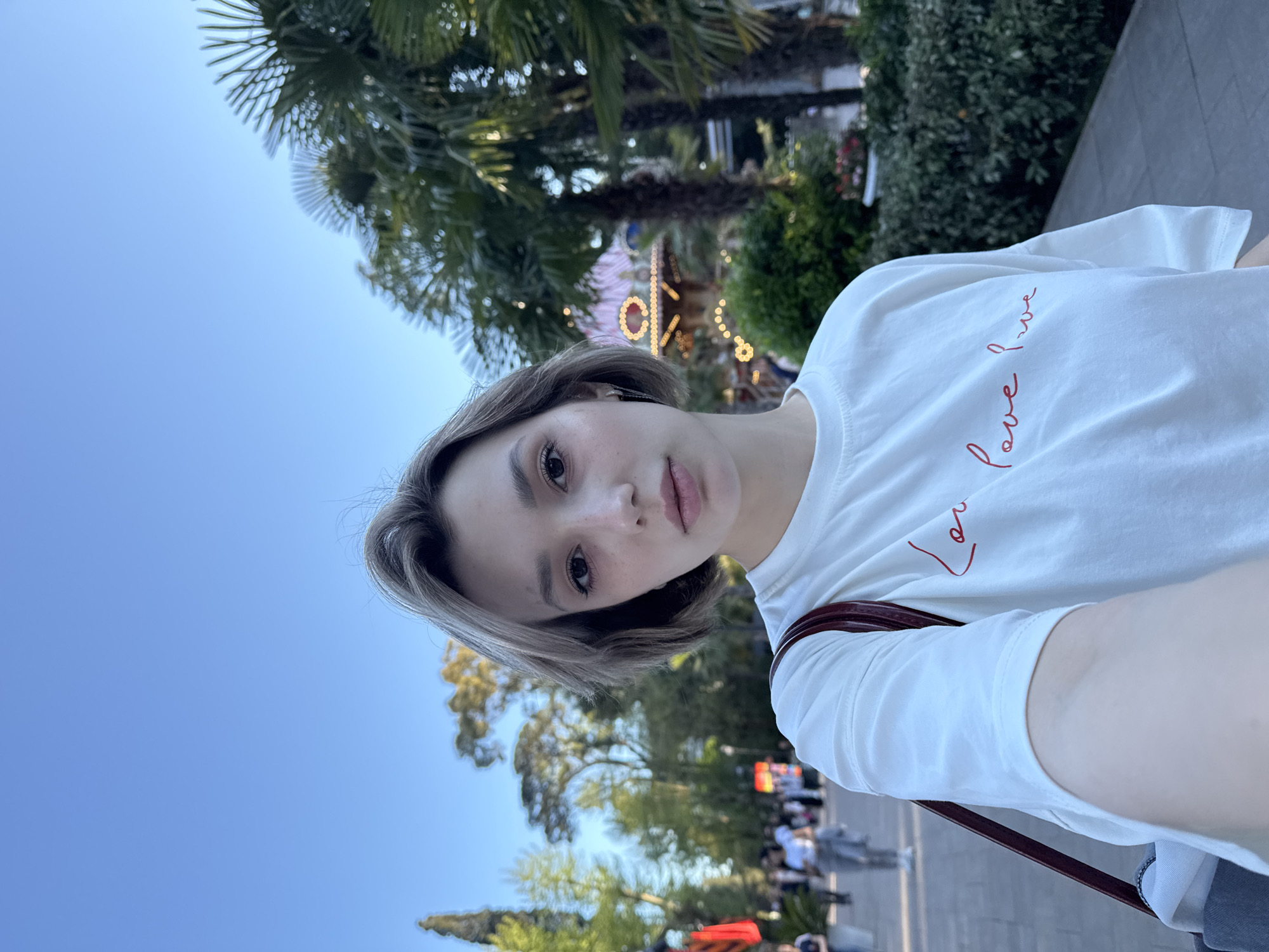 A woman is taking a selfie outside on what appears to be a city sidewalk. She has short, light brown hair with visible freckles on her face and is wearing a white T-shirt with a red script design. A brown strap from what seems to be a purse or bag is visible on her shoulder. Behind her, there are palm trees, green bushes, and other trees. In the background, a carousel with lights is partially visible, along with a crowd of people walking in the distance. The sky is blue and clear.