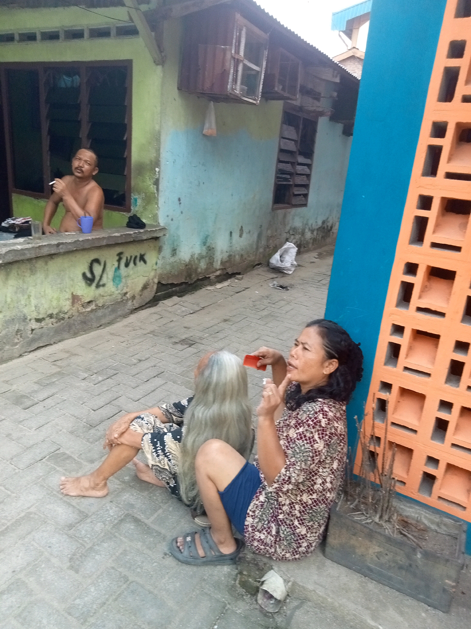 A topless man with a mustache is sitting with his back to a window, holding a cigarette in his right hand and a blue cup in front of him. He is in front of a faded green and blue painted building. To his left, graffiti reads "SL FVCK". Two women are sitting on the ground in front of the building. One woman, facing away from the camera, has long grey hair and is wearing a dark patterned dress. The other woman, facing the camera, has dark hair and is wearing a patterned shirt and blue shorts, holding a small orange object in her right hand. A tall orange brick structure is to her right. The ground is paved with grey bricks.