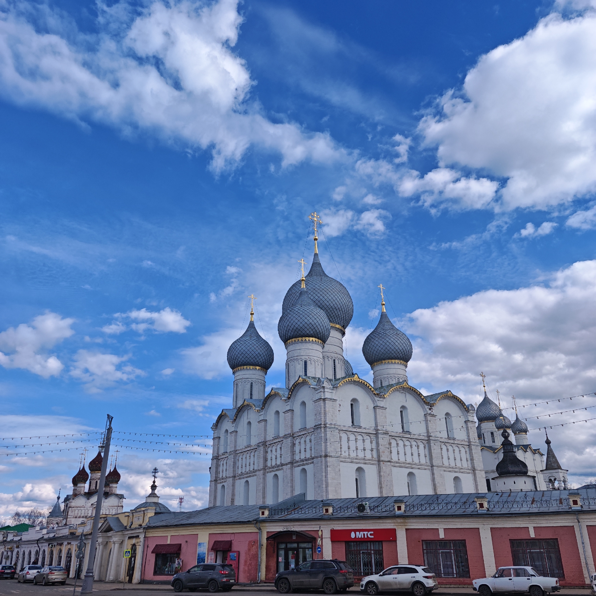 The image shows the Cathedral of the Assumption in Rostov, Russia, against a bright blue sky filled with scattered white clouds. The cathedral is a large white building with distinctive grey onion-shaped domes topped with golden crosses. The foreground features a row of brick buildings painted in various colors, including pink and red, with storefronts at street level. Cars are parked along the street in front of these buildings. The presence of an "MTC" sign suggests the location is in a commercial area. A street lamp and overhead wires are visible on the left side of the image.