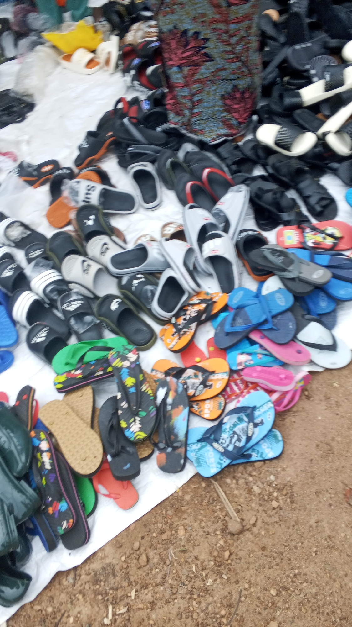 This image depicts an outdoor market stall in Nyita, Nigeria, displaying a large assortment of footwear. The primary items for sale are flip-flops and sandals, arranged haphazardly on a white tarp spread on the ground. Various colors and patterns are visible, including black, white, orange, blue, and multi-colored designs. Some of the footwear appears to be new, while others show signs of wear. In the background, a portion of a fabric garment with a floral pattern is visible, likely part of the stall's display or belonging to the vendor. The ground surrounding the stall is composed of dirt and small stones, indicating a typical market setting. There are no people clearly visible interacting with the merchandise, suggesting the image was taken from a distance or when the stall was unattended.