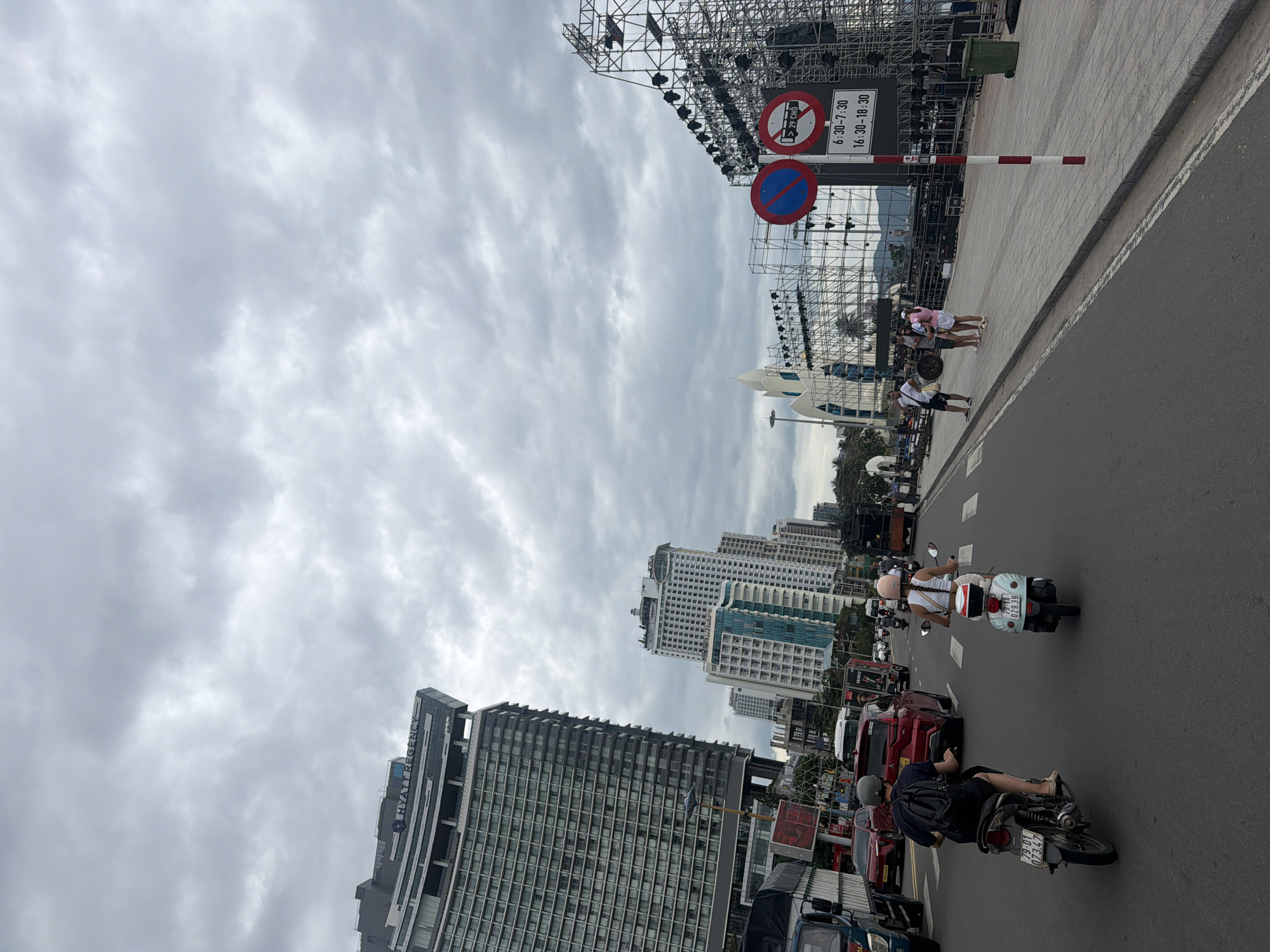 The image shows a street scene in Nha Trang, Vietnam, under a cloudy sky. On the left side of the street, a tall, modern building with the "Hyatt Regency" logo is visible, alongside other high-rise buildings. The street is busy with traffic, including cars, trucks, and motorcycles. In the foreground, two people are riding motorcycles. On the right side of the street, a large scaffolding structure, likely for an event or concert, is set up. Several traffic signs are present, including a "no parking" sign and a sign indicating restricted hours for vehicles. A group of people are standing on the sidewalk near the scaffolding. The overall environment suggests a bustling urban area with ongoing activities or preparations.