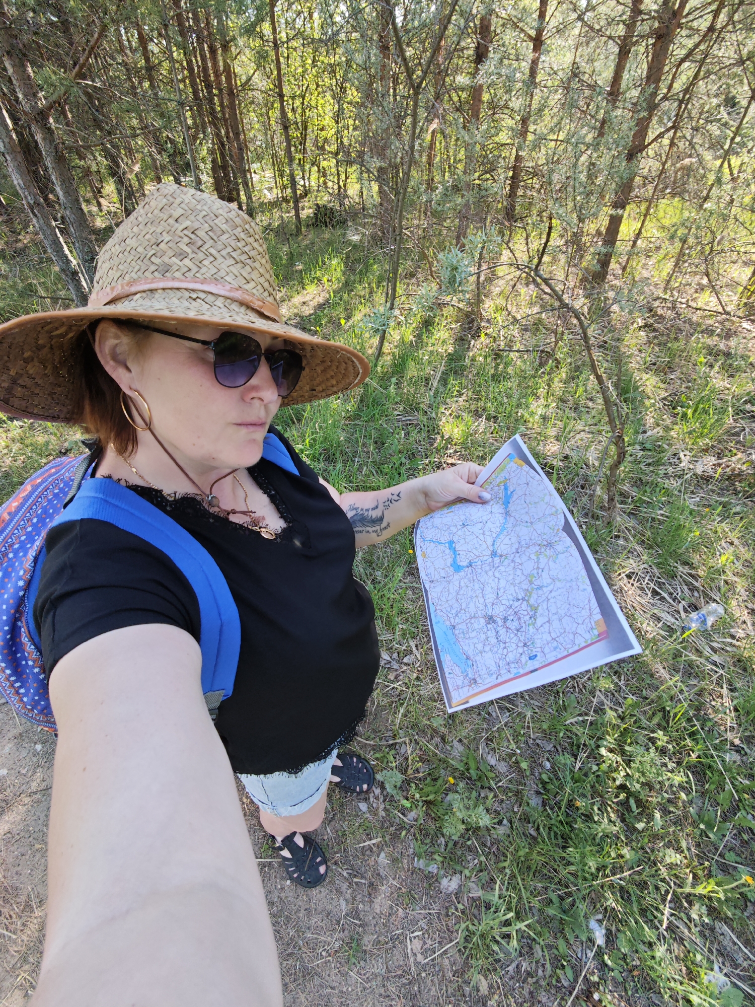 A woman stands in a wooded area, holding a map. The woman is wearing a straw hat with a pink band, sunglasses, a black shirt with lace detailing, denim shorts, and black sandals. She has a blue backpack on her back, gold hoop earrings, and a necklace. Tattoos are visible on her arm. She is holding a printed map in her right hand, which appears to show a network of roads and bodies of water.
The environment consists of a wooded area with trees, grass, and vegetation on the ground. A dirt path is visible.
The image was taken at eye-level.