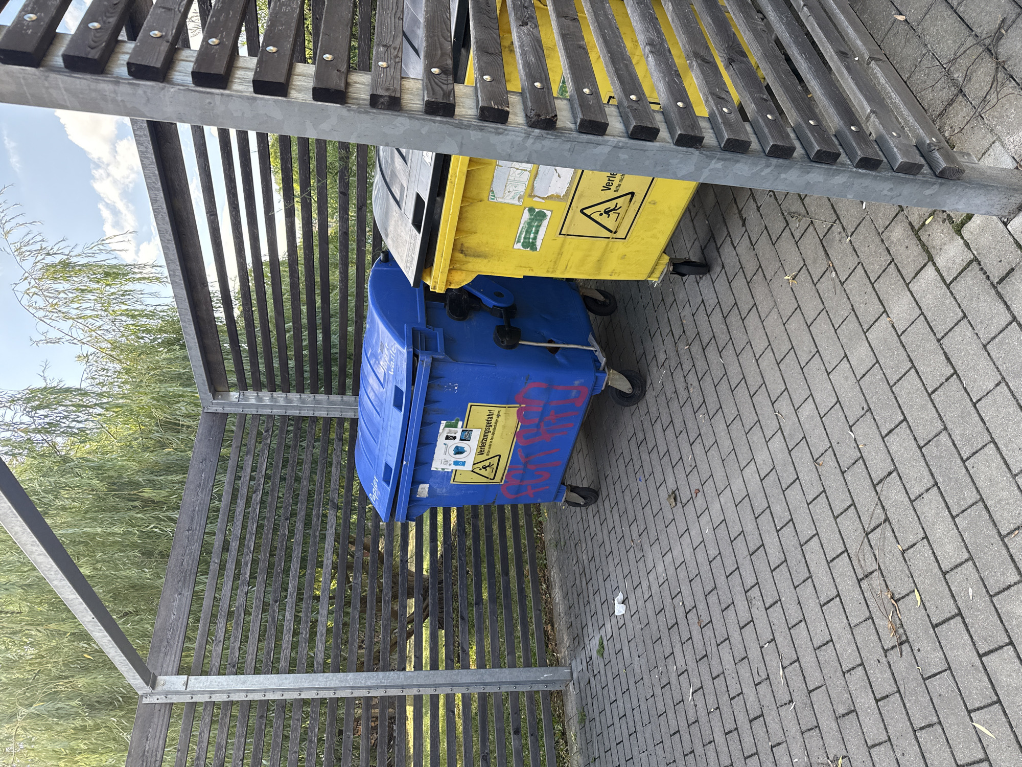 In Bad Blankenburg, Germany, a fenced-in outdoor area contains two large, wheeled trash receptacles. A blue container, labeled "Papier," is positioned to the left, adorned with red graffiti. To its right is a yellow container, also with a lid, which bears a warning sticker depicting a figure tripping. The ground is paved with grey brickwork, and scattered leaves and a small piece of white paper are visible on the pavement. A wooden slatted fence encloses the area, supported by metal posts, with a backdrop of green trees and a blue sky with scattered clouds.