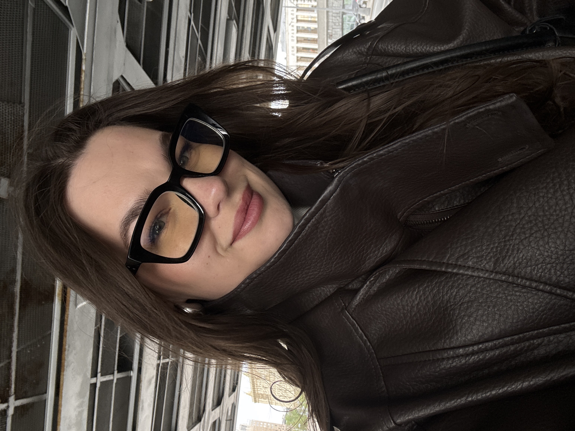A medium shot depicts a woman with long, dark brown hair and fair skin. She is wearing black-framed glasses and a dark brown leather jacket. Her facial expression is pleasant, with a slight smile. The background features metal bars and, out of focus, cityscape elements suggestive of Moscow, Russia. The image's orientation is rotated slightly to the left.