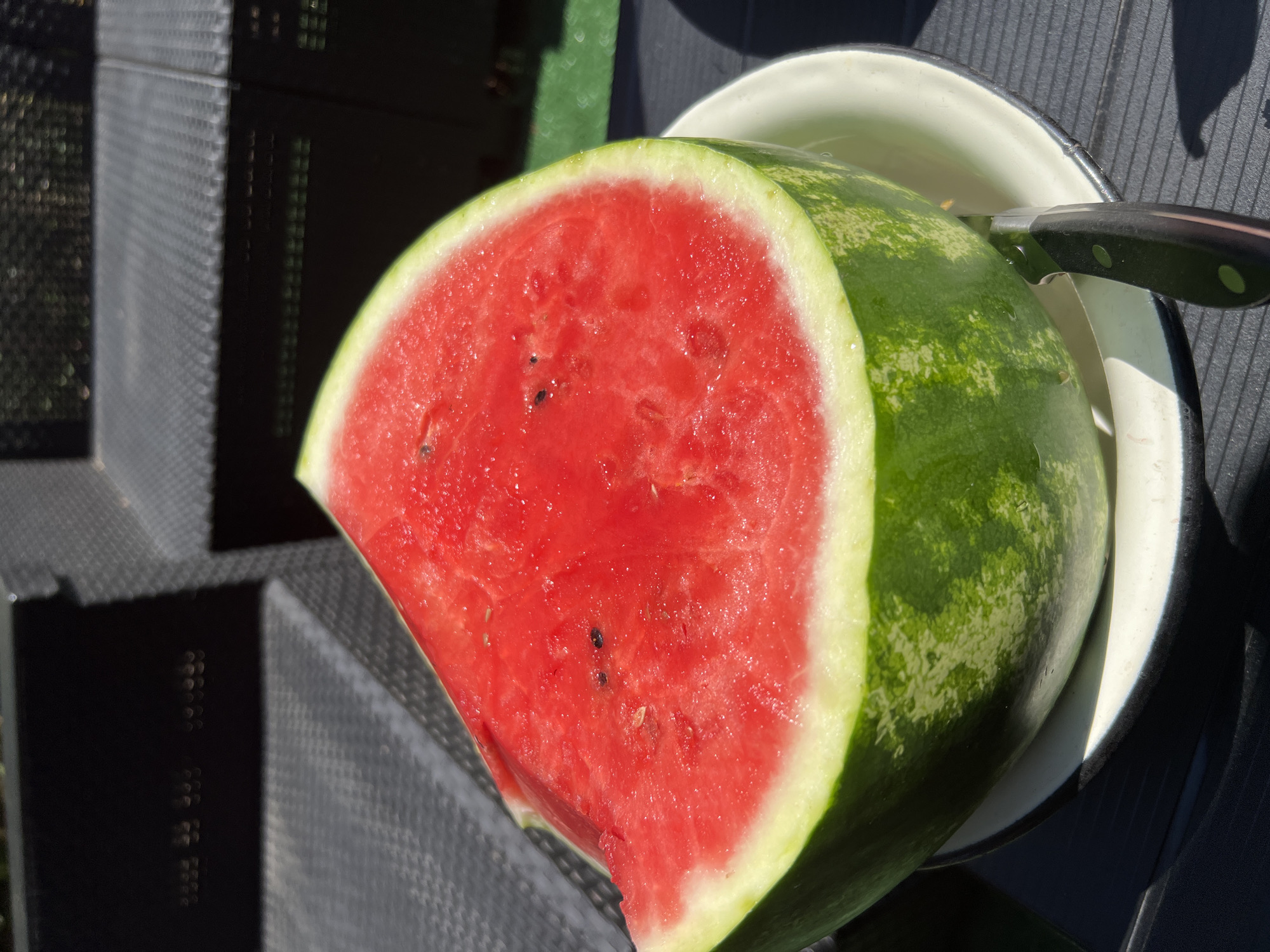 A cut watermelon rests on a white plate with a black rim, which is situated on a dark, textured surface, possibly a table or outdoor furniture. A knife with a black handle is partially inserted into the watermelon. The watermelon is bright red inside with visible seeds and has a thick green rind with lighter green variegations. The background shows dark, textured surfaces, likely outdoor furniture, and out-of-focus greenery, suggesting an outdoor setting. The lighting indicates it is daytime, with strong sunlight casting shadows. There are no people visible in the image. The location is not specified in the image or its provided context.