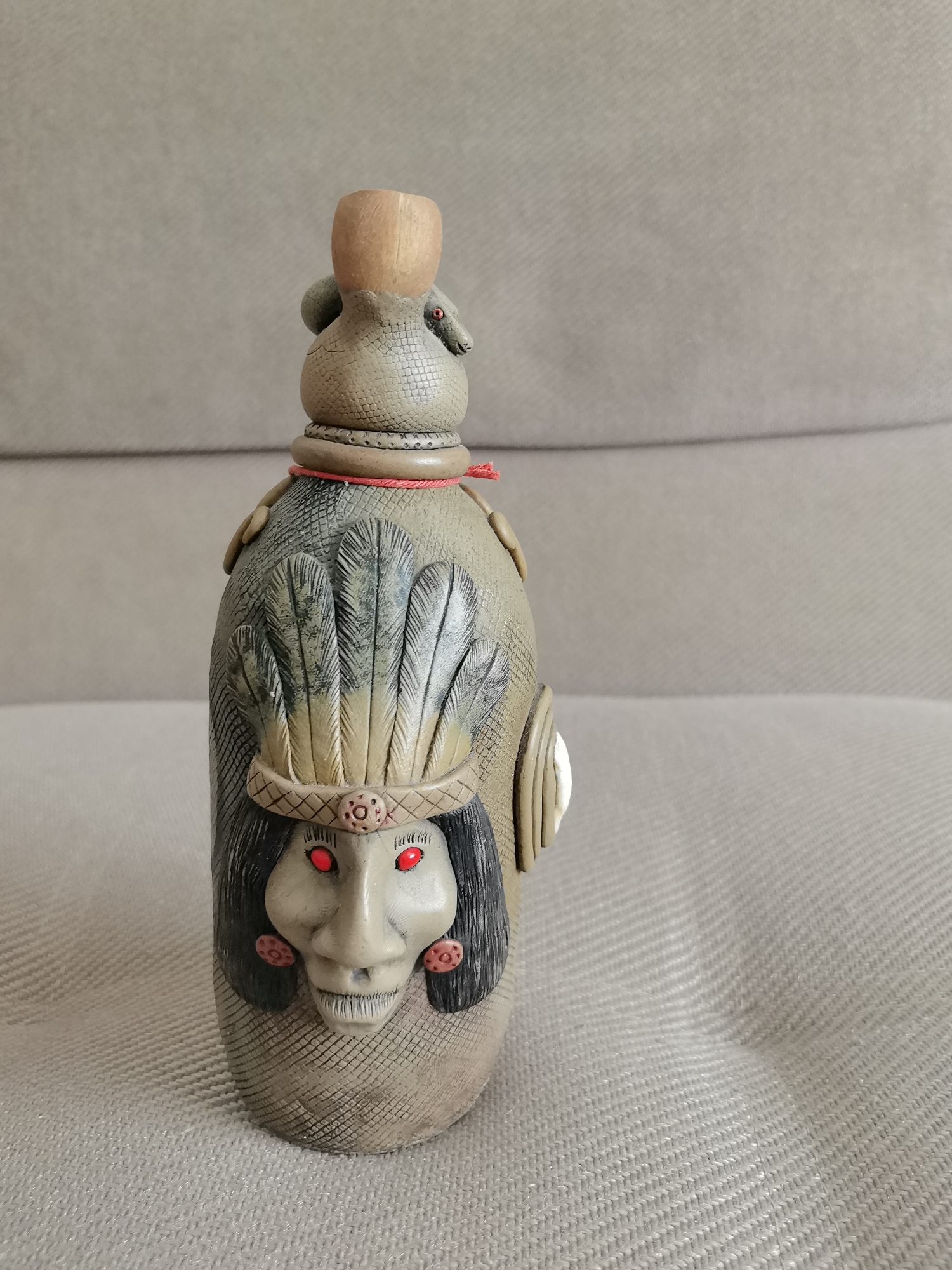 A ceramic bottle shaped like a stylized Native American figure sits on a textured gray fabric surface, possibly a sofa. The bottle is tan with dark gray detailing, featuring a face with bright red eyes, black hair, and an elaborate headdress of feathers. A thin red cord wraps around the neck of the bottle. The bottle has a wooden stopper. The background is a plain, textured gray surface. There is no indication of the city of Oryol, Russia, in the image.