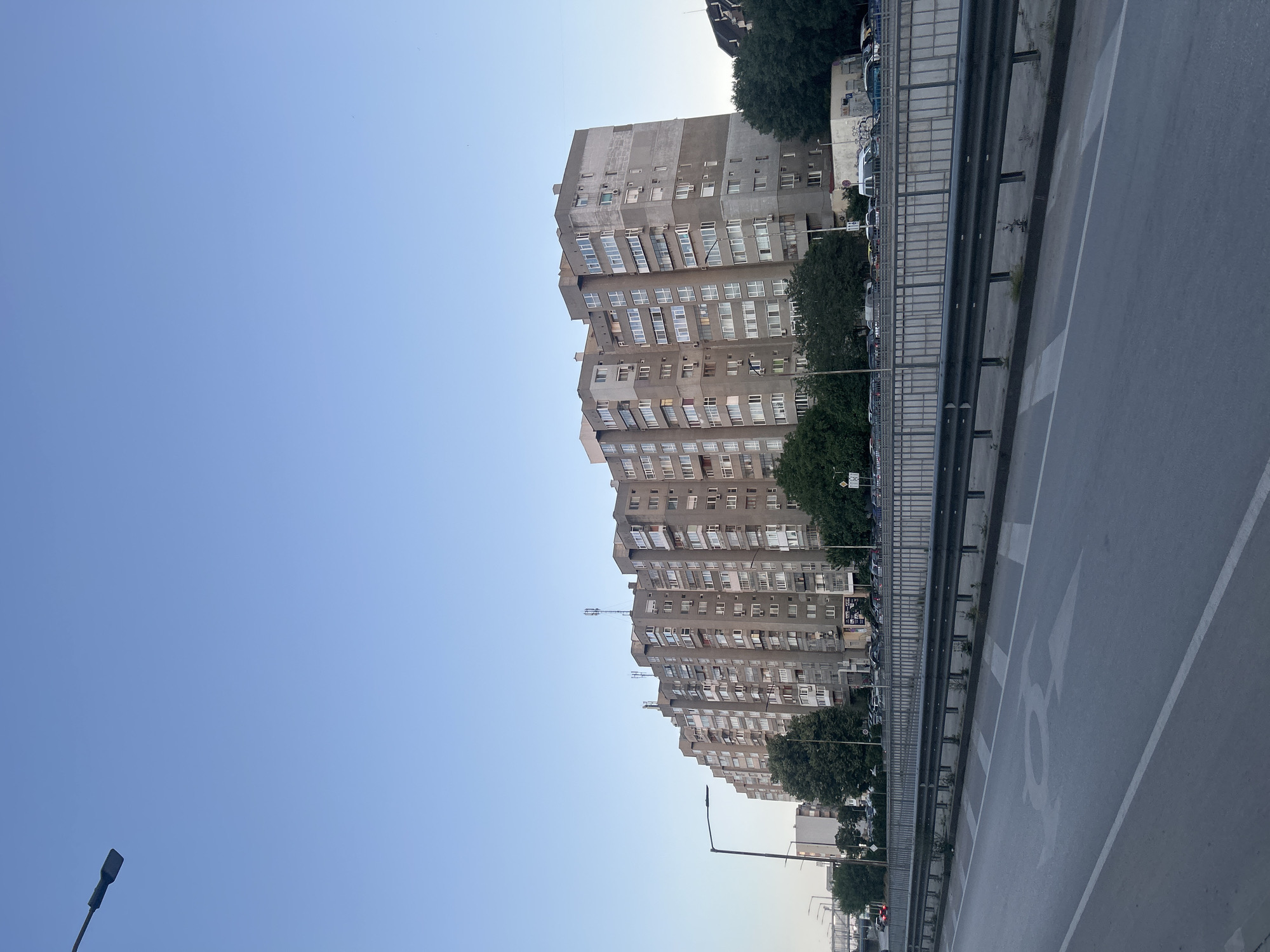 The image depicts a wide, multi-lane road in Varna, Bulgaria, with a prominent, long, Brutalist-style apartment building dominating the background. The building, constructed of grey concrete, features a staggered facade with numerous balconies and windows. The sky is clear and light blue, indicating either early morning or late afternoon. Along the side of the road, there is a metal guardrail separating the roadway from a slightly elevated area where trees and parked cars are visible. Further beyond the apartment building, other structures and more trees can be seen. The overall impression is an urban residential area with a large infrastructure.