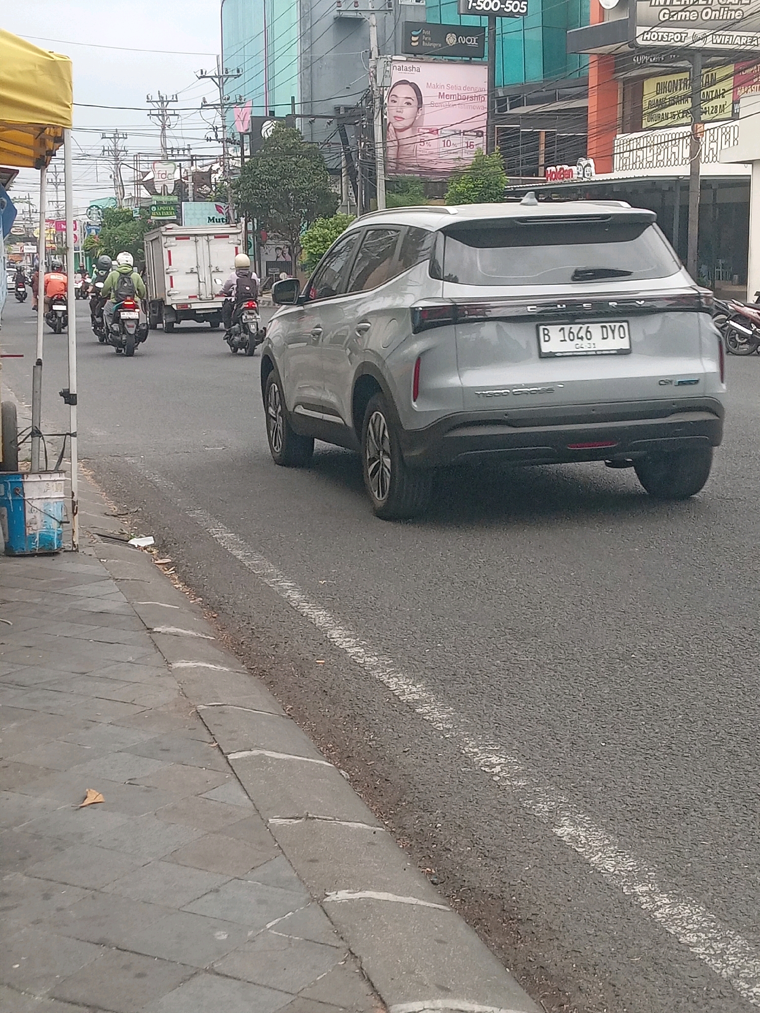 The image captures a street scene in Depok, Indonesia, likely on a Tuesday, April 31st (implied by the license plate date). A silver Chery Tiggo Cross is the prominent subject, facing away from the camera and moving down a paved road. Its license plate, "B 1646 DYO," is clearly visible. Several motorcycles, with riders wearing helmets and some with backpacks, are also on the road, some ahead and some beside the car. One motorcycle in the foreground has a rider wearing a green jacket, and another has a rider in white. In the background, a white box truck is visible. Buildings line the street, including one with a large green glass facade and signage that includes "APOTEK" and "MUTI." Another building has a prominent advertisement featuring a woman's face and text in Indonesian, likely for a membership program. Utility poles and wires are abundant overhead. The sidewalk is paved with square tiles, and a yellow awning extends from a stall on the left side of the image. A blue and white painted metal drum with a tire on top is positioned on the sidewalk near the stall. The overall environment appears to be an urban street with moderate traffic.