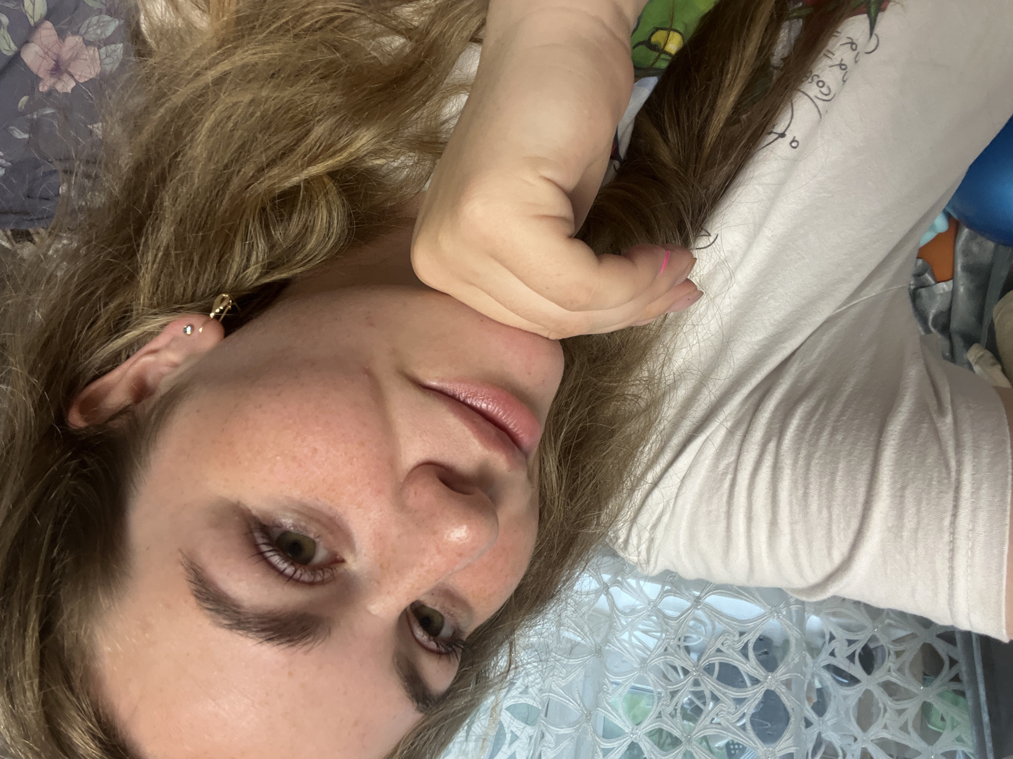 A woman with long, light brown hair is lying down, looking at the camera. She has fair skin, freckles, and green eyes. Her left hand is positioned under her chin. She is wearing a light-colored t-shirt with a print on it. An earring is visible in her left ear. The background includes a floral pattern fabric and a white lattice-like structure. The location is not discernible from the image.