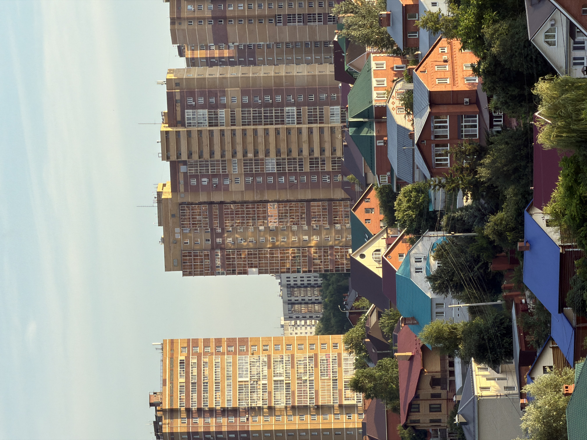 This is a photograph of a cityscape in Kazan, Russia, taken from a high vantage point. The image is oriented vertically, with the sky occupying the left side and the buildings and trees on the right. In the background, several tall, multi-story apartment buildings with a grid-like pattern of windows dominate the skyline. These buildings appear to be made of concrete and brick, with variations in color from light brown to darker reddish-brown.
In the foreground and middle ground, a neighborhood of smaller houses is visible. These houses have pitched roofs in various colors, including green, blue, and maroon, and are surrounded by lush green trees. Some of the houses are made of brick, while others have wooden facades. The contrast between the imposing apartment blocks and the more intimate residential area suggests a mix of urban development.
The time of day appears to be late afternoon or early evening, as indicated by the warm, soft lighting that casts gentle shadows. The sky is a pale blue with a few wispy clouds, suggesting fair weather. The overall impression is one of a densely populated urban environment with a blend of architectural styles. There is no visible text in the image.