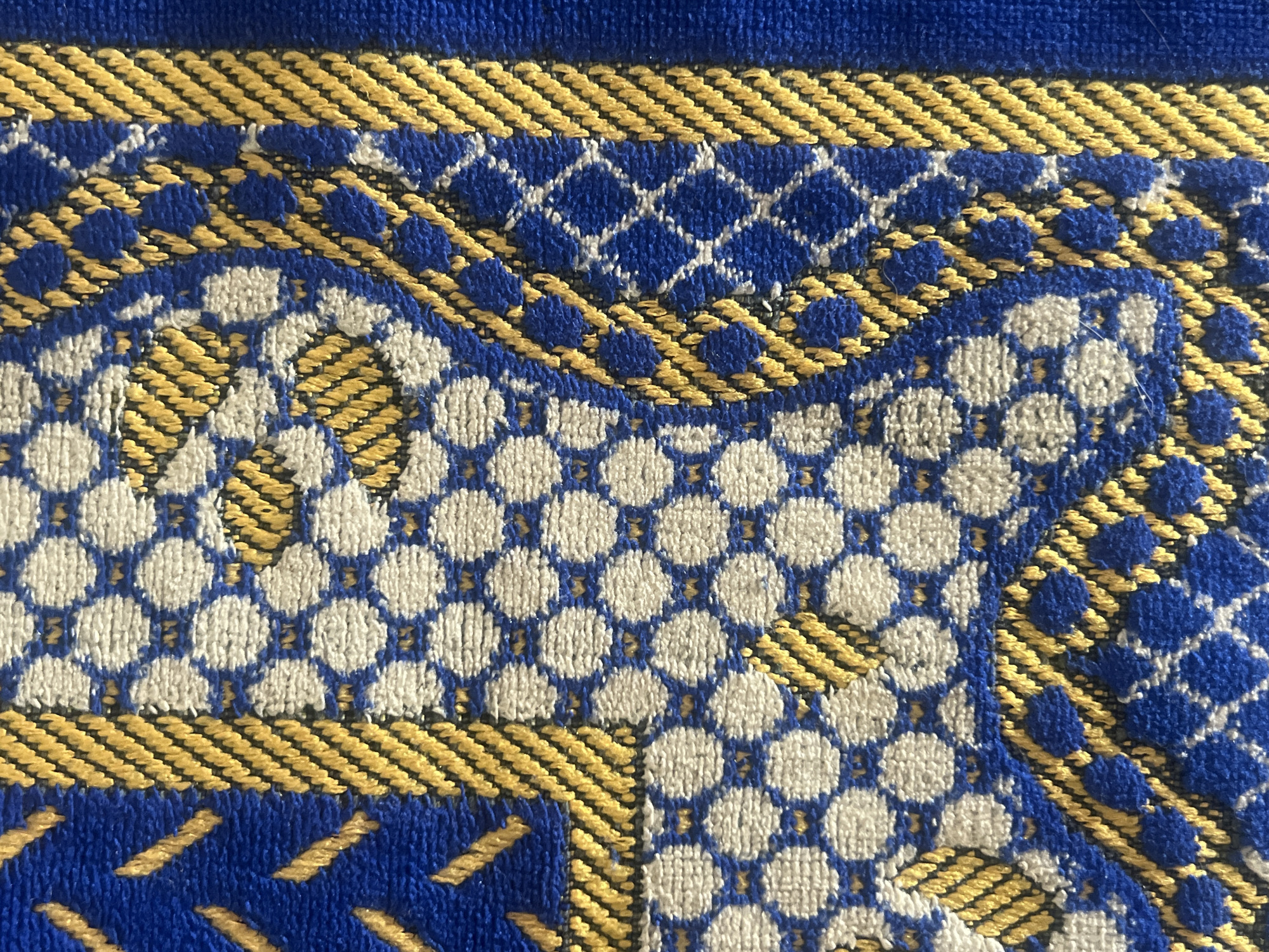 This is a close-up photograph of a woven textile, located in Casablanca, Morocco. The fabric exhibits a plush, textured pile, predominantly featuring deep blue, golden yellow, and off-white colors.
The textile's design is intricate, combining geometric and curvilinear patterns:
1. **Off-white sections:** These areas are patterned with small, regularly spaced blue circular dots, forming a dense hexagonal grid. Some of these sections also contain isolated golden yellow dashes or partial outlines.
2. **Golden yellow diagonal stripes:** Strips of tightly packed, parallel golden yellow lines are set against a solid deep blue background, creating borders and framing elements within the larger design. These stripes run diagonally.
3. **Golden yellow diamond grid:** A lattice pattern of interconnected golden yellow diamonds is visible on a deep blue background in various sections.
4. **Curvilinear motifs:** Flowing, stylized shapes are outlined in golden yellow against the deep blue background, sometimes interacting with the off-white dotted areas.
The overall appearance suggests a robust, ornamental fabric, likely a rug or heavy upholstery. The threads show visible texture and slight variations in pile, indicative of its woven construction. No people, actions, or specific environmental details beyond the textile itself are discernible in the image.