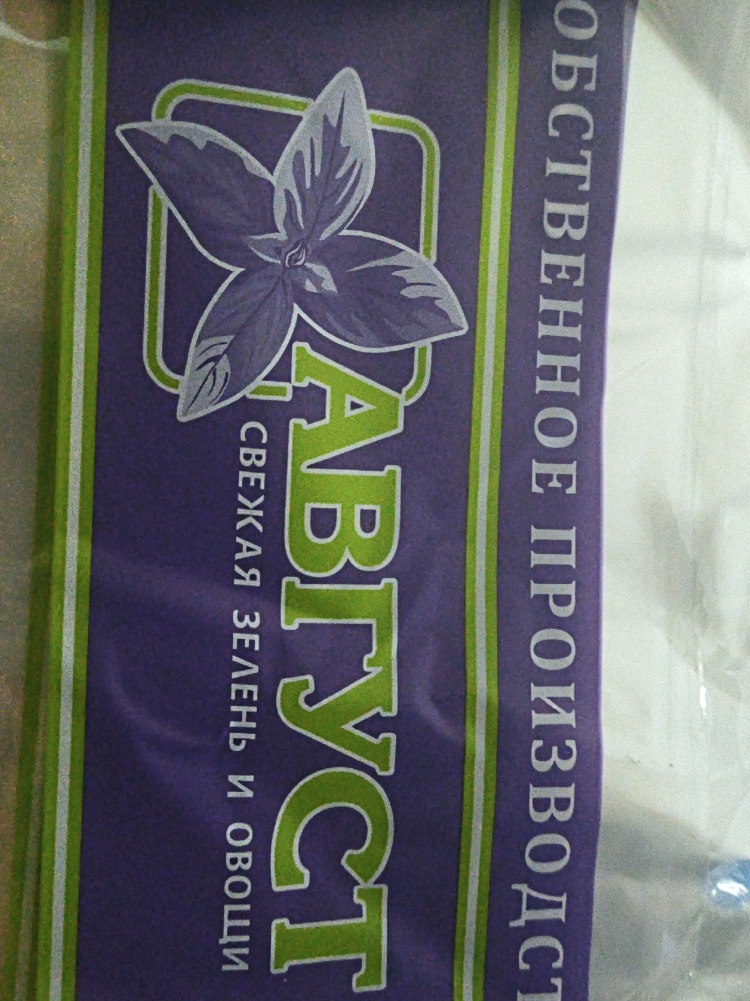 The image shows a close-up of a purple package with green and white stripes. The package features a stylized illustration of a basil plant with the word "АВГУСТ" (August) written in large, green, stylized letters. Below this, in smaller white text, are the words "СВЕЖАЯ ЗЕЛЕНЬ И ОВОЩИ" (Fresh greens and vegetables). To the right of the main text, written vertically in white, is the word "ОБЩЕСТВЕННОЕ ПРОИЗВОДСТ" (Public production). The lighting suggests it is indoors, possibly in a retail or storage setting, but there are no other objects or people visible to provide context about its specific location within Uzhevka, Russia.