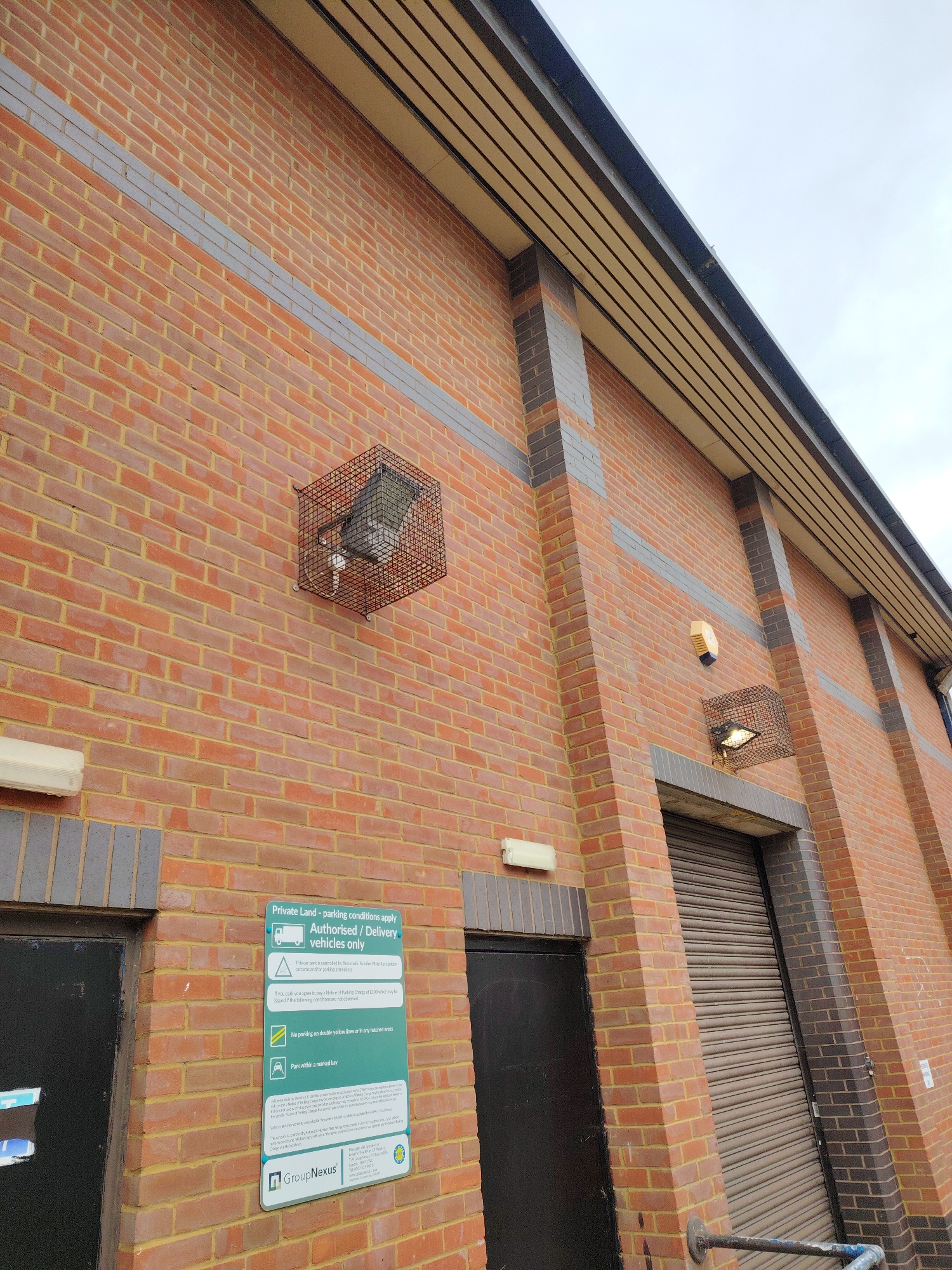 This image depicts the exterior wall of a commercial or industrial building in Peterborough, United Kingdom, under an overcast sky. The wall is predominantly constructed from orange-red bricks with yellow mortar. Horizontal bands of lighter grey bricks run across the wall, and darker grey/brown bricks form vertical pillars and lintels around openings.
Mounted on the upper left section of the wall is a large outdoor floodlight enclosed within a black wire mesh cage. A similar floodlight, also caged, is positioned above a large roller shutter door on the right side of the image. Above this right-side floodlight, a cream-colored security alarm box is affixed to the wall. Below the level of the upper grey brick band, two cream-colored rectangular light fixtures are visible; one is to the left of a black pedestrian door, and the other is to the left of the large roller shutter door.
Between the pedestrian door on the left and the roller shutter door on the right, a vertical green and white informational sign is mounted. The sign details "Private Land - parking conditions apply" for "Authorised / Delivery vehicles only," stating that the car park is controlled by "Automatic Number Plate Recognition cameras and/or parking attendants" and managed by "GroupNexus."
The pedestrian door on the left is plain black. The large opening on the right is covered by a closed, dark brown/grey corrugated roller shutter door. A dark, possibly weathered, metal handrail extends diagonally from the bottom-right corner of the image, appearing to be fixed near the roller shutter door. No people or vehicles are present in the scene.