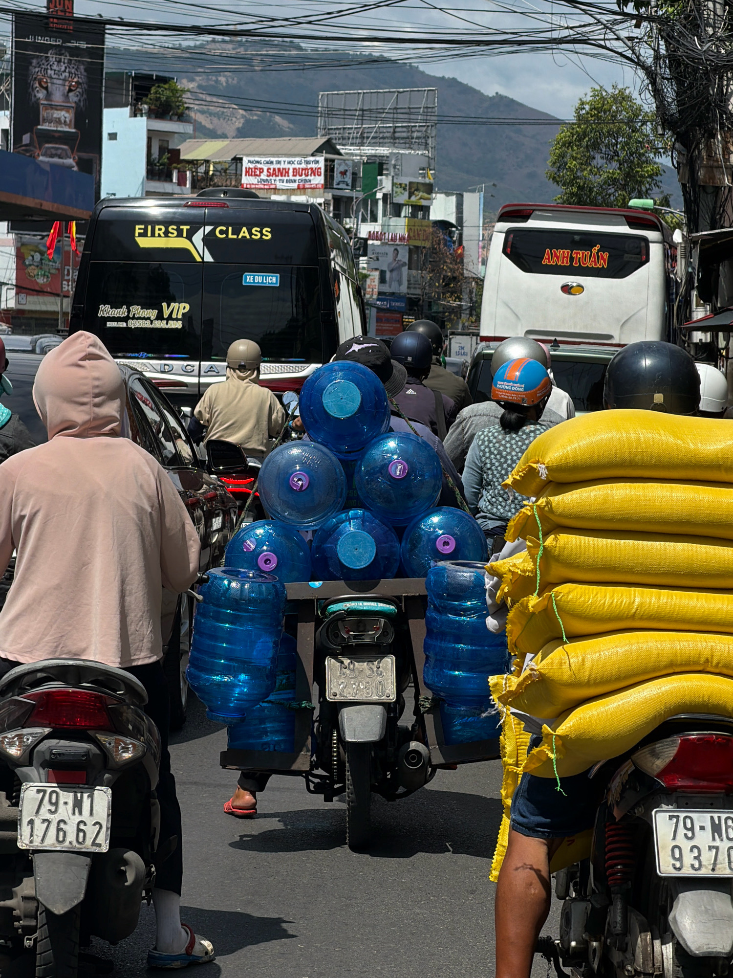 This is a street scene in Nha Trang, Vietnam, capturing a moment of traffic congestion. The primary focus is on a motorcycle laden with multiple large blue water jugs, stacked precariously and secured with green string. To the right of this motorcycle, another motorcycle is heavily stacked with large yellow sacks, also bound with green string. In the background, several buses are visible. One black bus on the left has "FIRST CLASS" emblazoned on its side in yellow lettering, along with Vietnamese text. A white bus on the right has red Vietnamese text that reads "ANH TUẤN". People on motorcycles, many wearing helmets, are interspersed throughout the scene. One person in the foreground is wearing a light pink hoodie. The environment suggests a busy urban setting with overhead electrical wires and various buildings lining the street. The sky is overcast, and the lighting indicates it is daytime.