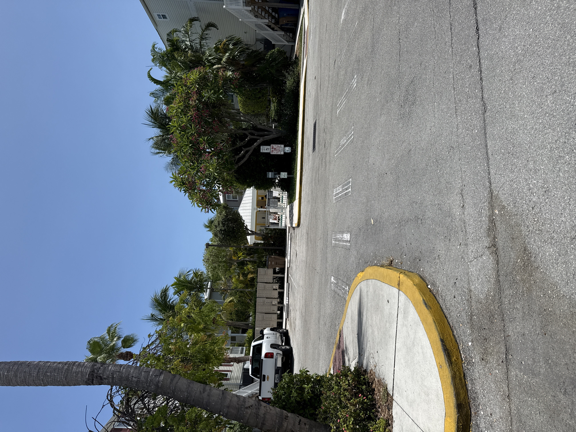 A white pickup truck is parked in a lot near a row of mailboxes. The lot is paved and there are white lines painted on the ground to designate parking spaces. A sidewalk with a yellow curb is visible on the left side of the image. In the background, there are buildings with palm trees and other tropical vegetation. The sky is clear and blue. The location is Stock Island, United States of America.